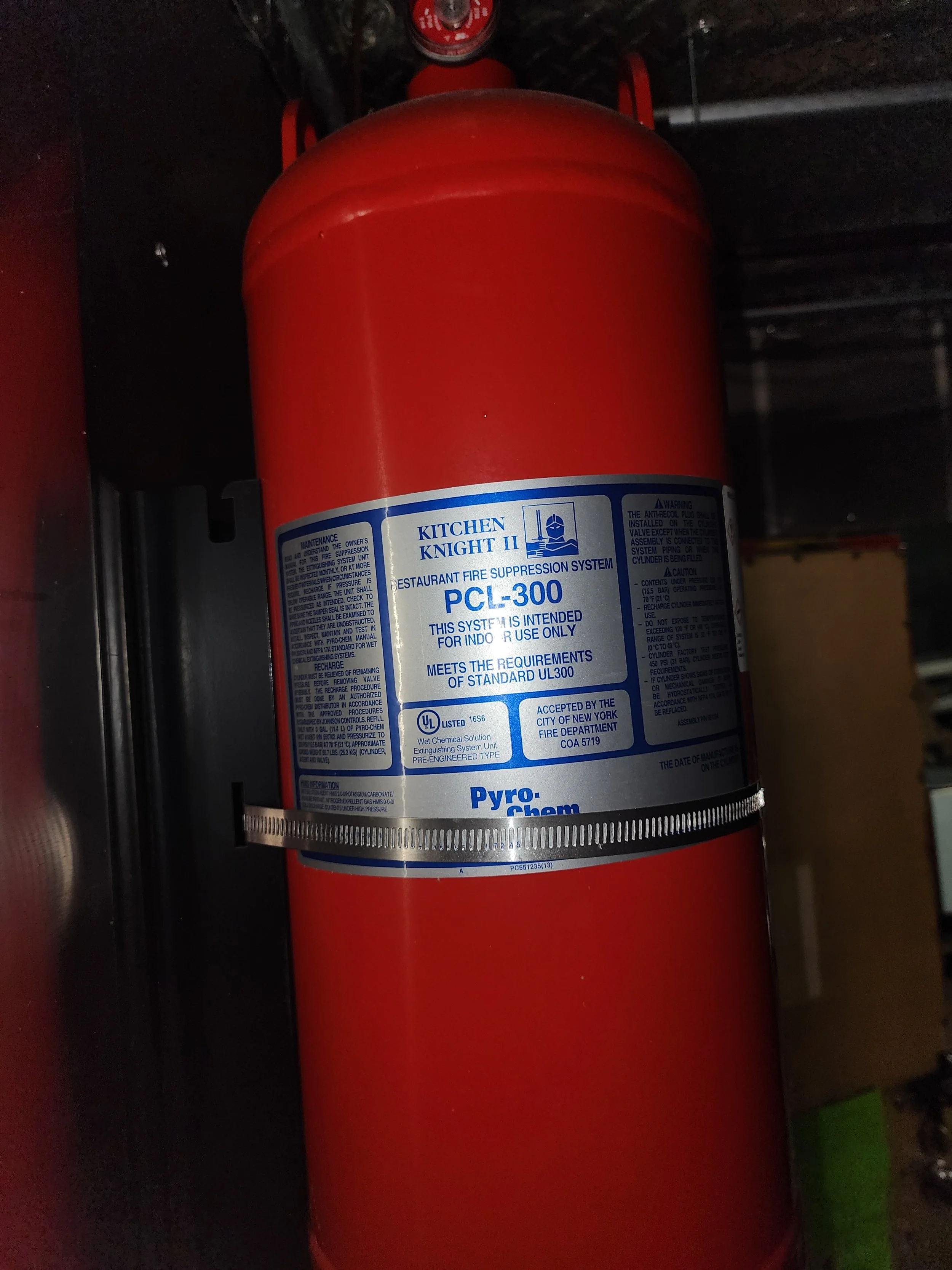 A red Kitchen Knight II fire suppression system cylinder labeled PCI-300, installed in a kitchen or similar setting.
