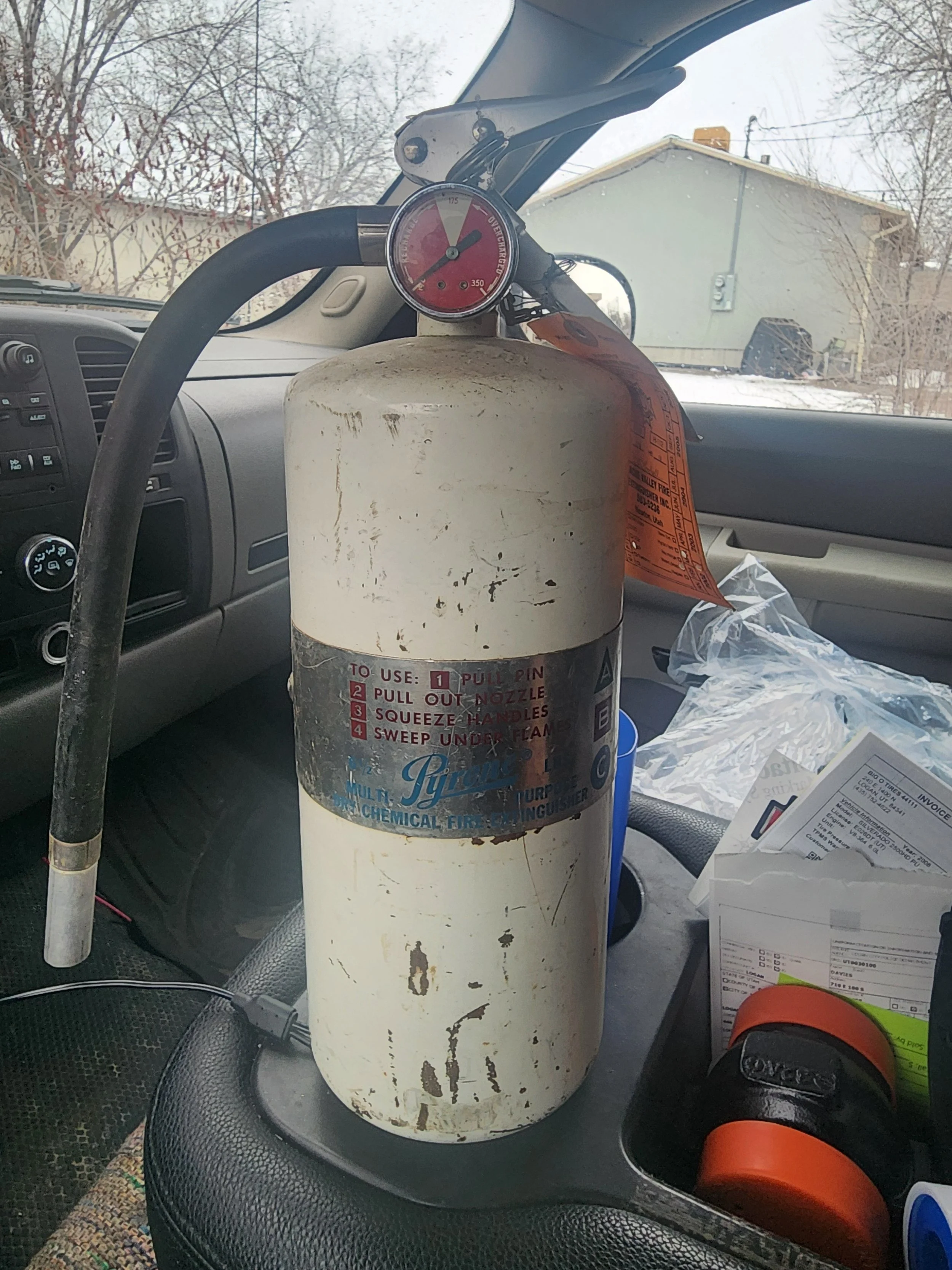 A fire extinguisher placed on a vehicle dashboard with a pressure gauge on top, surrounded by papers and an orange and black object, with a outside view of a house and trees through the windshield.