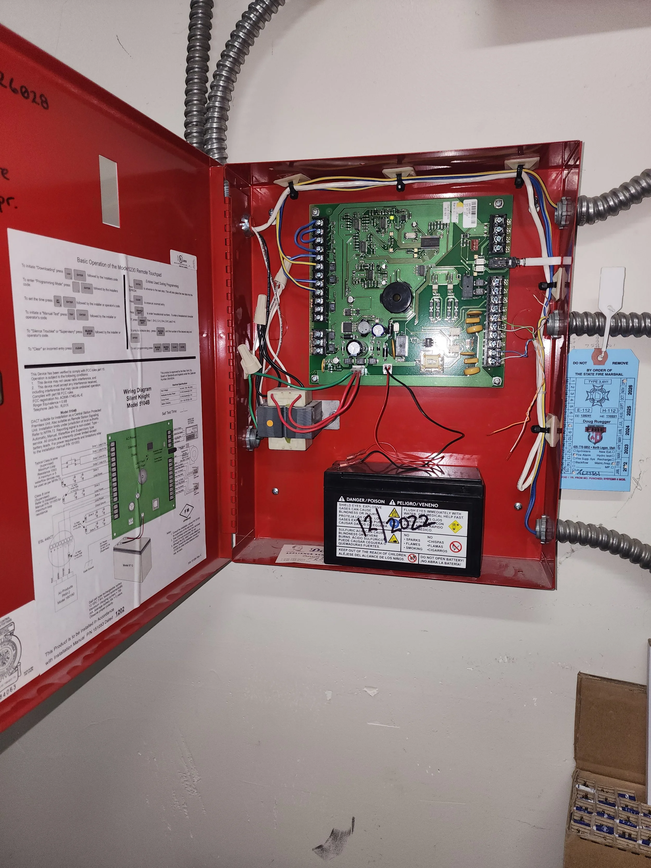 Interior view of a red fire alarm control box with its cover open, revealing a green circuit board, black battery, and various wiring.