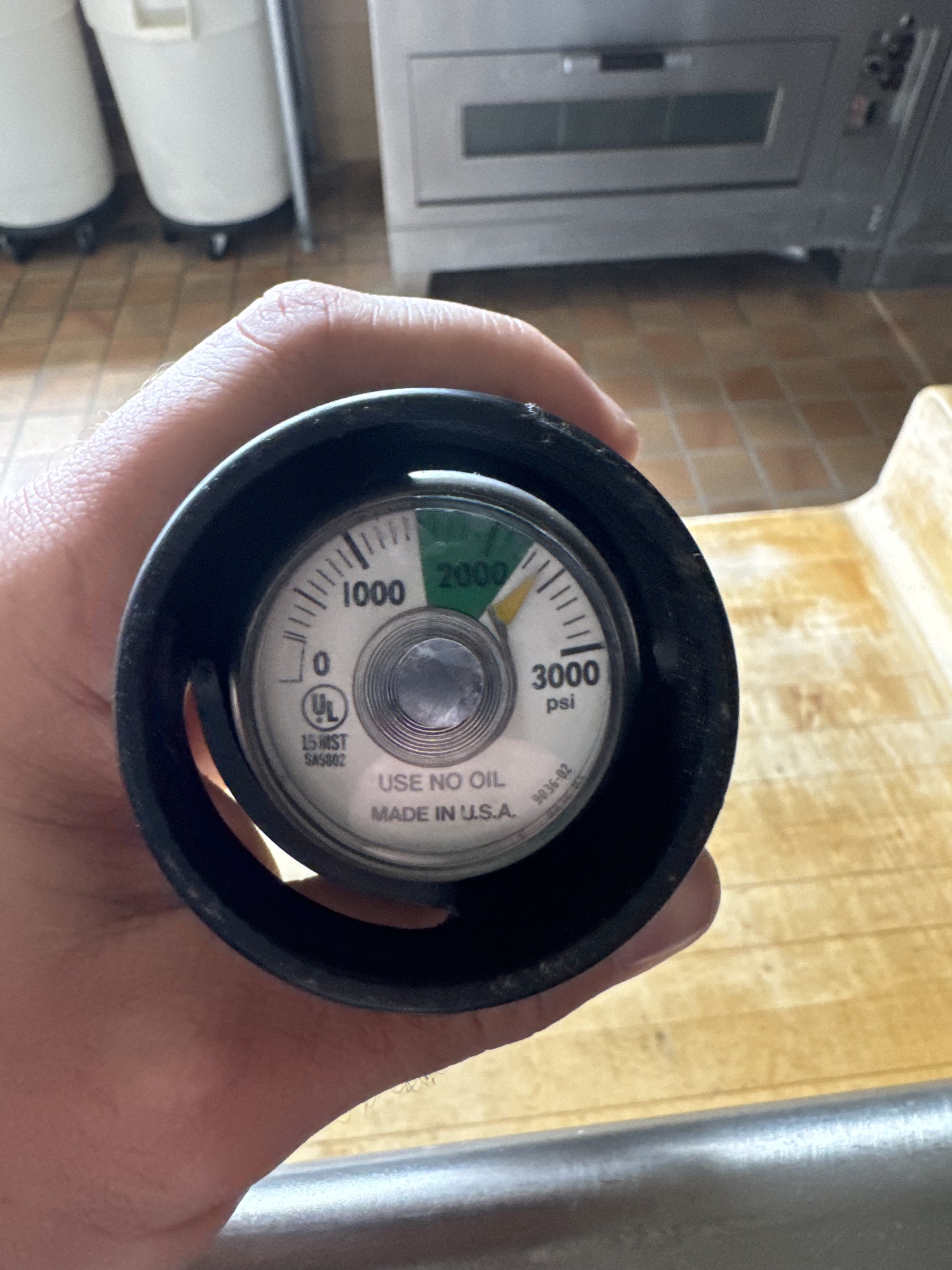 Hand holding a pressure gauge with a green zone between 2000 and 3000 psi, indicating pressure in pounds per square inch, with text on the gauge reading "USE NO OIL" and "MADE IN U.S.A." in a kitchen setting.