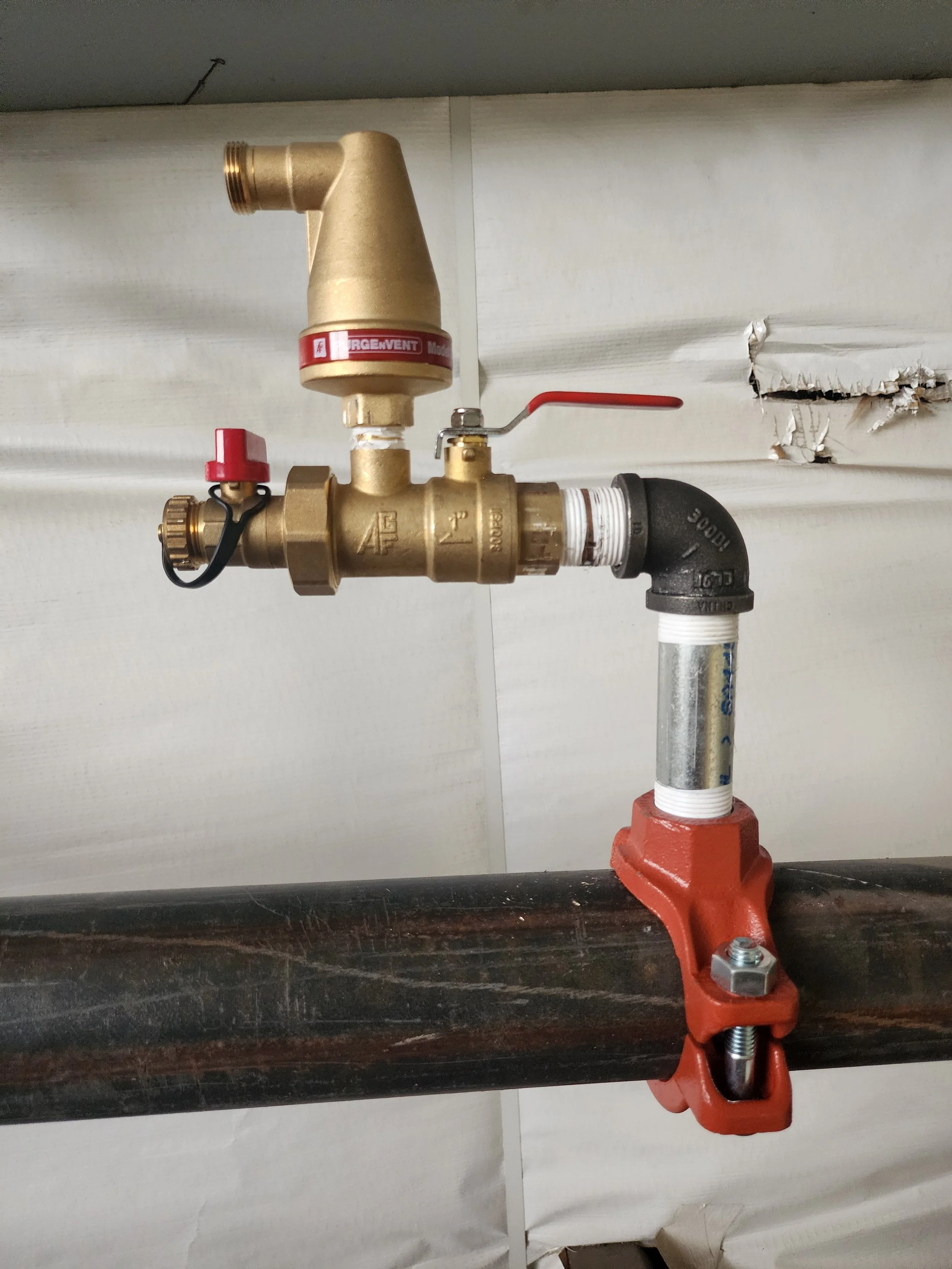 A plumbing valve assembly with brass and black pipes, a red handle, and a black elbow joint, mounted on a large dark pipe, with an unfinished wall background.