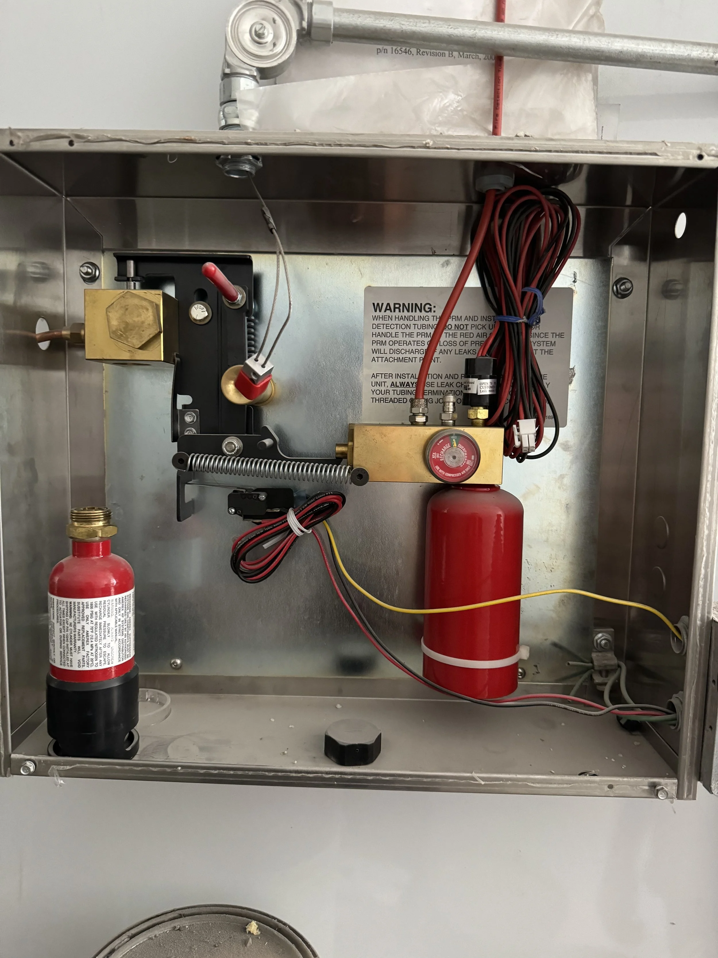 Inside a metal enclosure with electrical components, wires, a red cylinder, and a warning label, likely part of a fire suppression system.