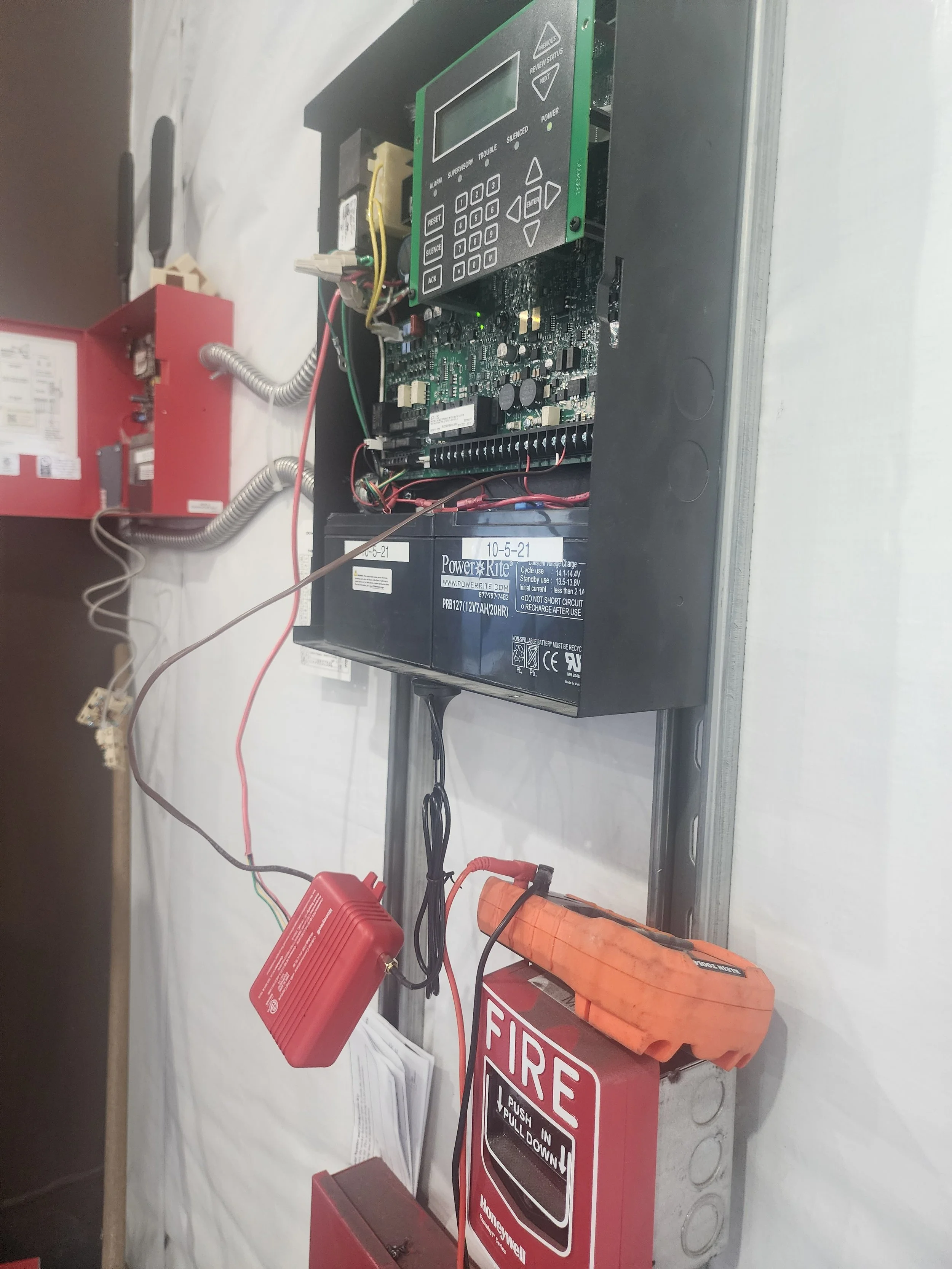 Electrical panel with circuit board, wiring, and a large battery, mounted on a wall with a fire alarm box below.