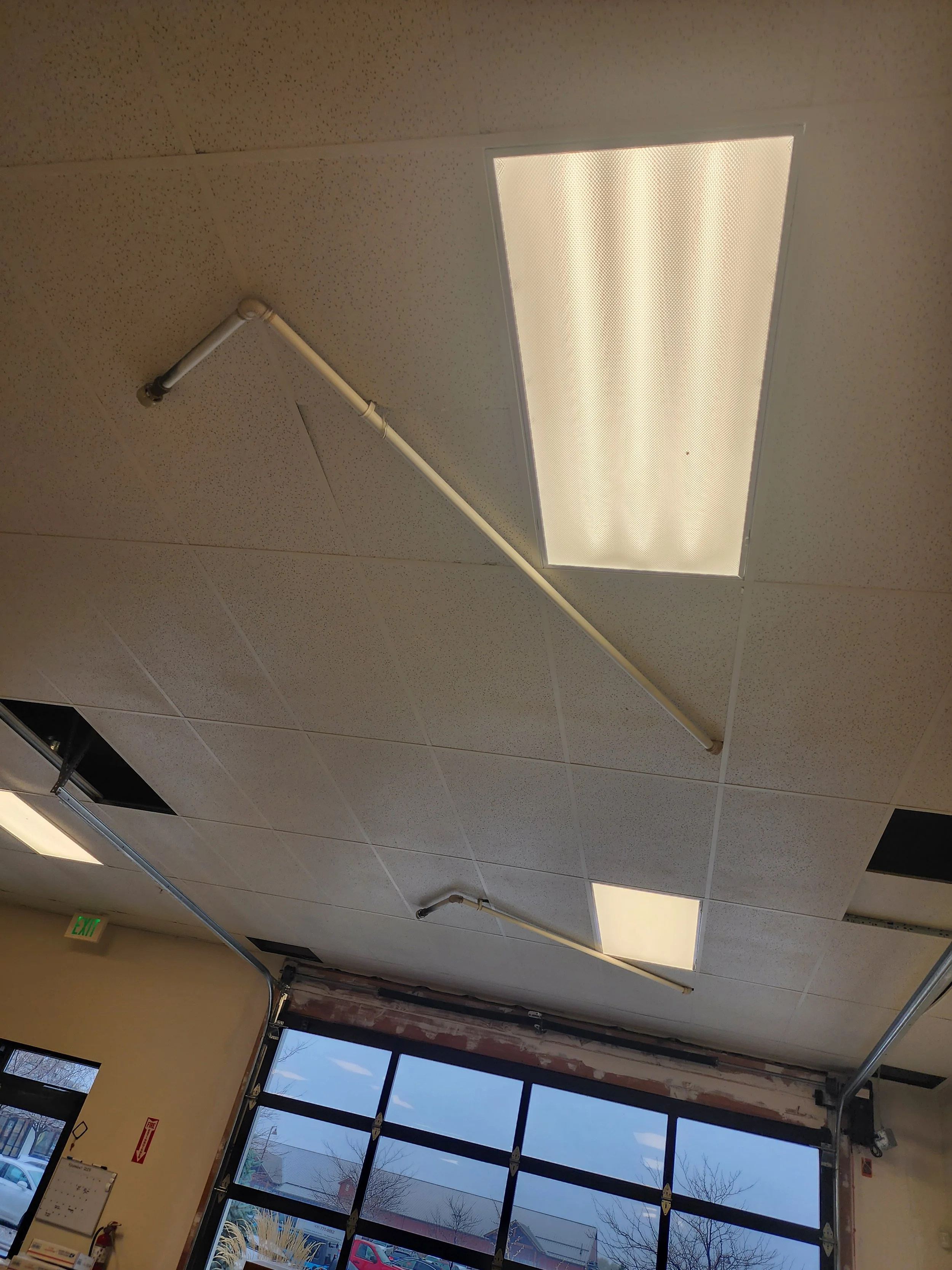 Ceiling of an indoor space with fluorescent light panels and white pipes, with a view of an open garage door and windows showing cars outside.