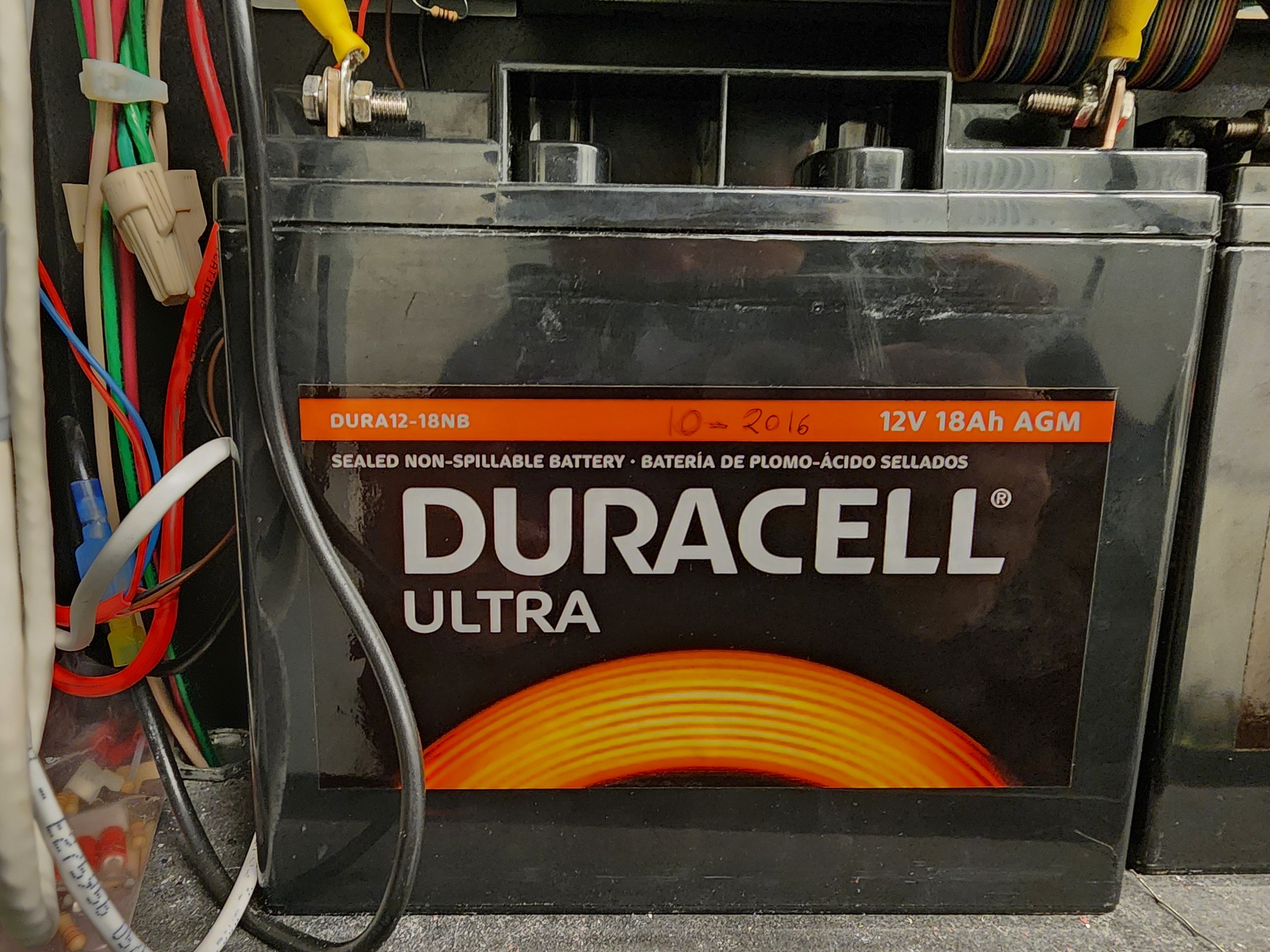 A Duracell Ultra 12V 18Ah sealed lead-acid battery with wires and electrical components nearby.