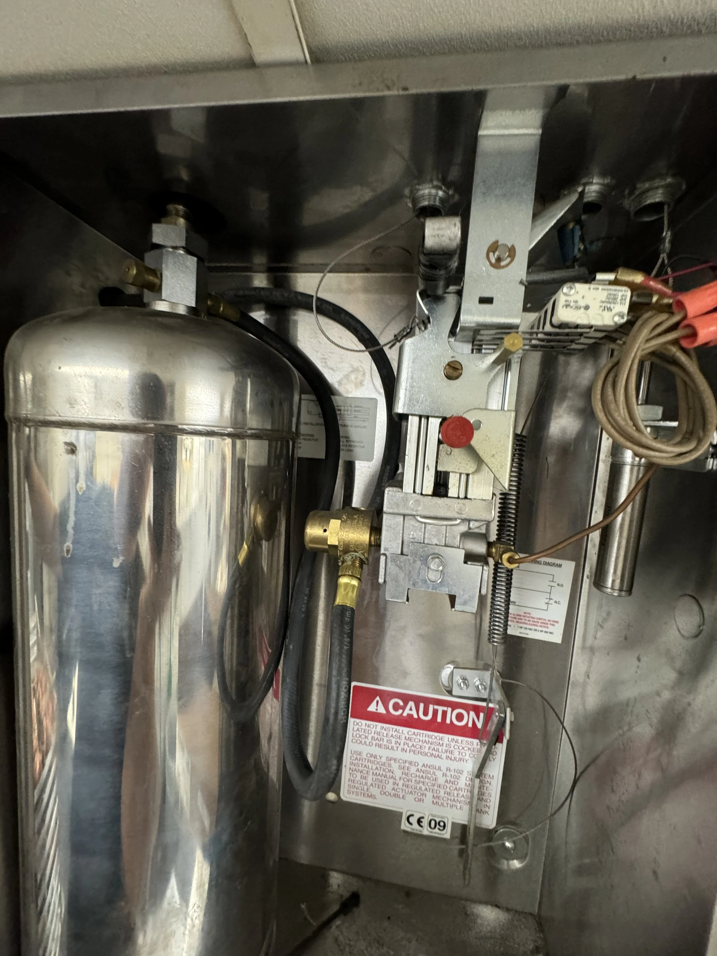 Close-up view of a mechanical device inside a metallic enclosure, including a large cylindrical silver component, various metal brackets, wires, and colorful wiring with warning labels.