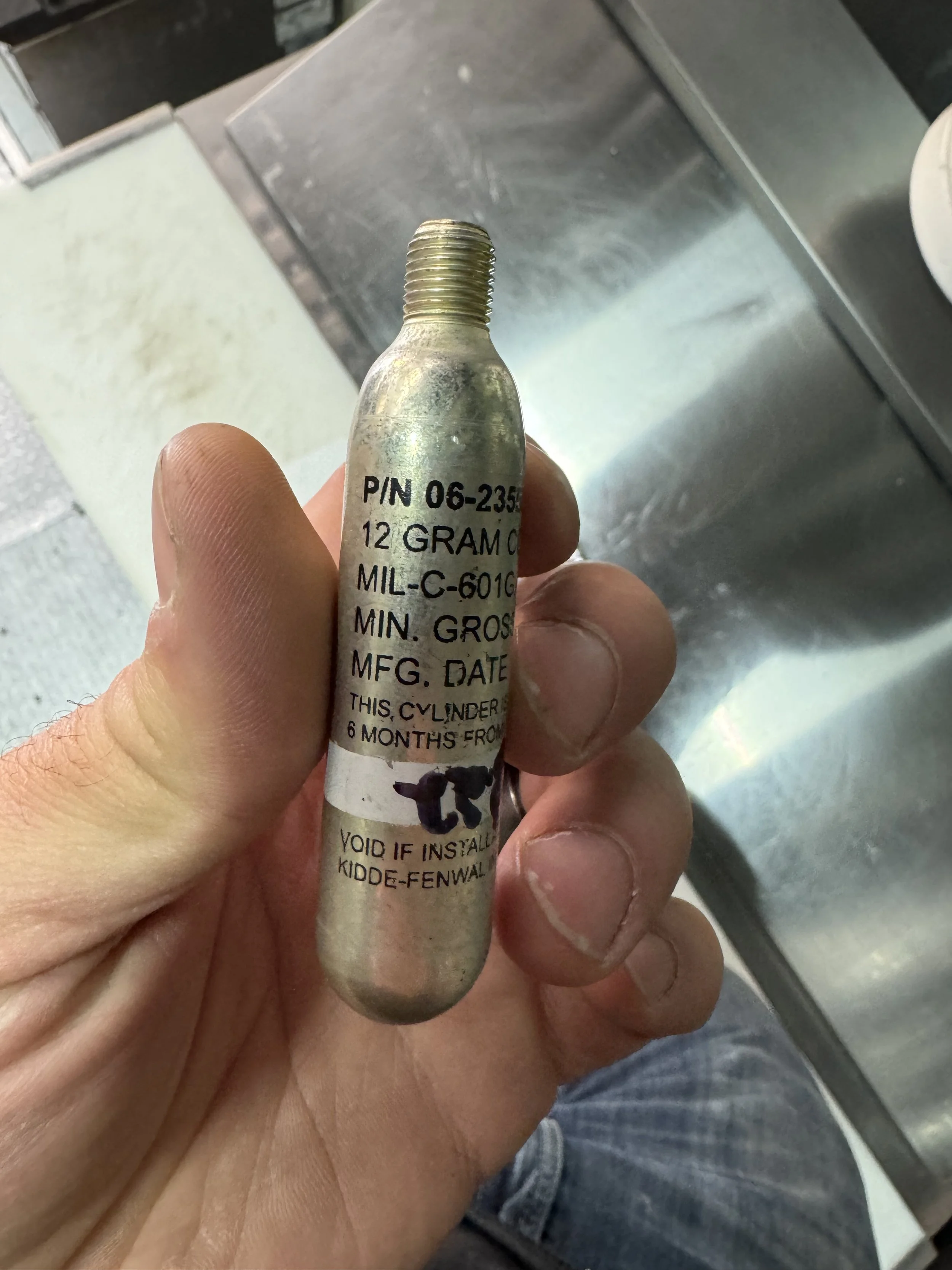 A person holding a small metal cylinder with printed technical information, including part number, weight, and manufacturing details, in a stainless steel kitchen environment.