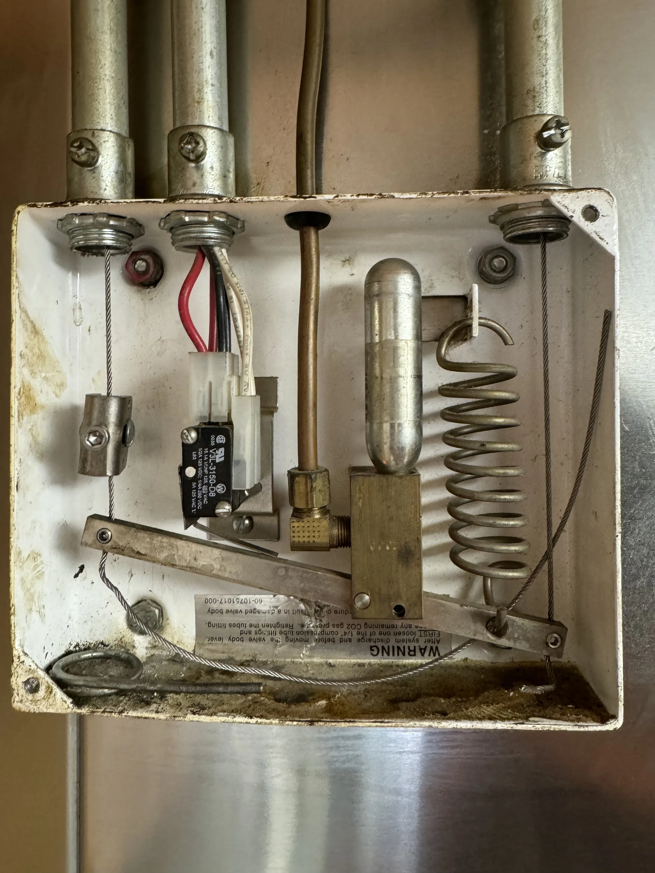 Inside a metal electrical box showing wires, a relay, a sensor, and a spring, with some dirt and corrosion at the bottom.