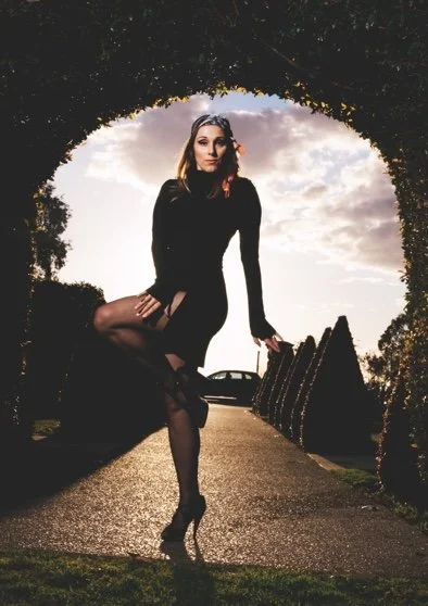 A woman in a black dress and high heels sitting on a bench outdoors during sunset, framed by trees and with a car in the background.