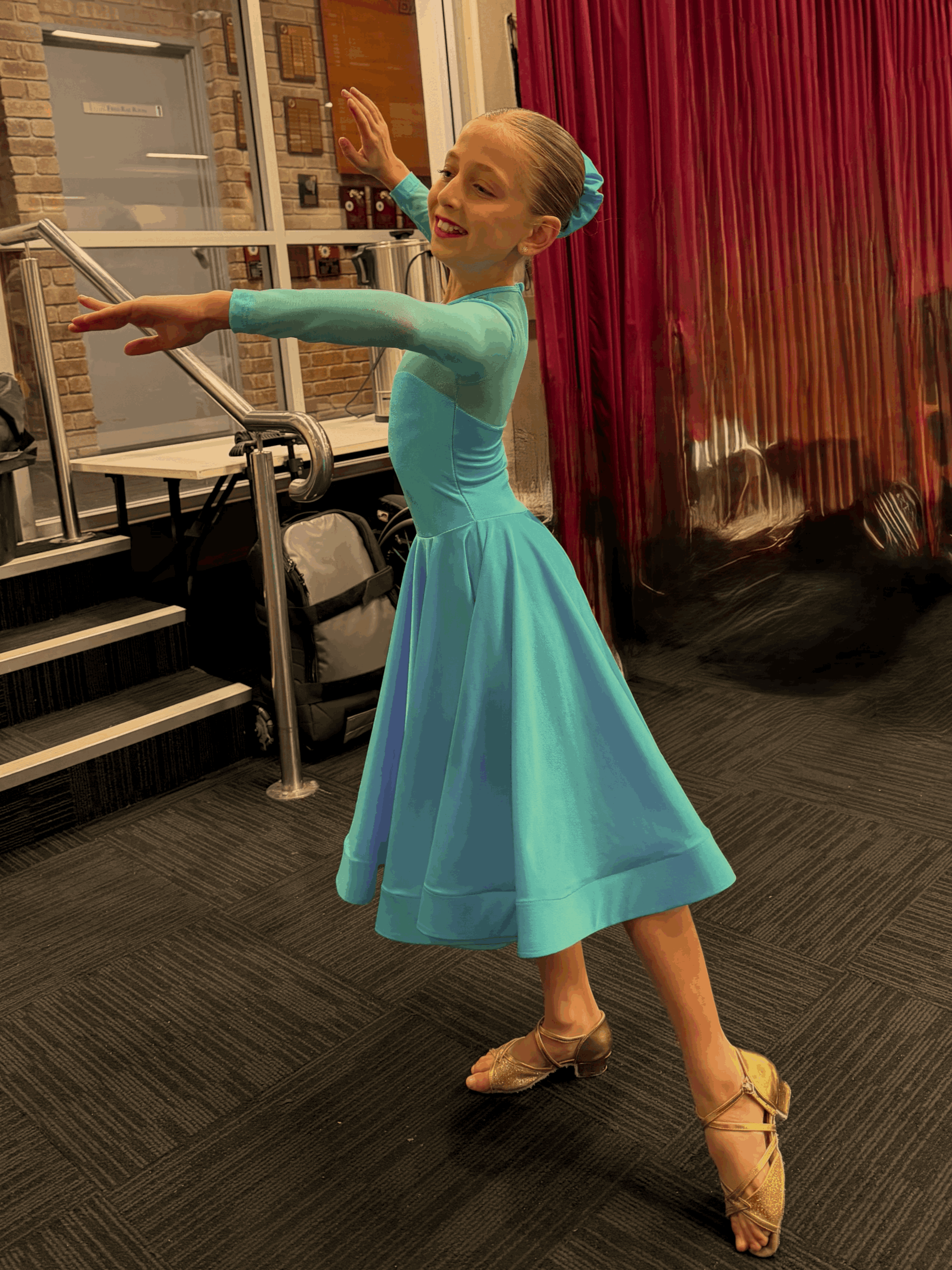 A young girl in a turquoise dance costume with a matching hair accessory is dancing on a gray carpeted floor. She is wearing tan ballroom dance shoes and is striking a dance pose with one arm extended and the other bent at her side. She is smiling with her eyes closed, and the background features brick walls, large windows, and red curtains.