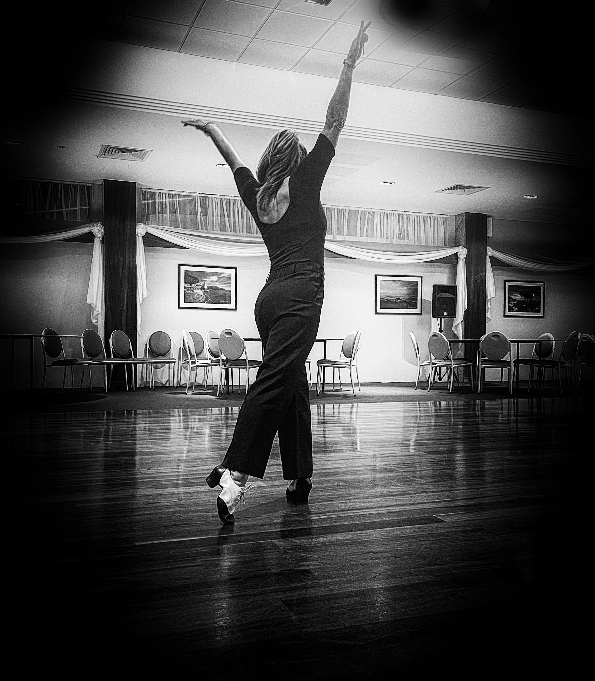 A woman dancing in a ballroom or dance hall with her arms raised above her head and one leg slightly bent. She is wearing a fitted black dress and high-heeled shoes. The background shows framed pictures on the wall, draped curtains, and rows of chairs, with a speaker on the right side of the image.