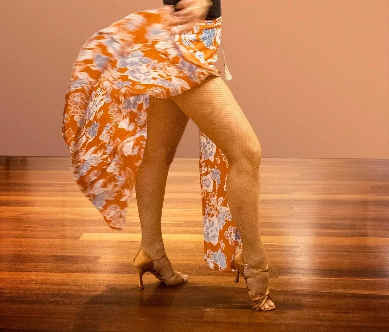 A person dancing in a floral orange and white dress, fishnet stockings, and high heels, standing against a plain wall and wooden floor.