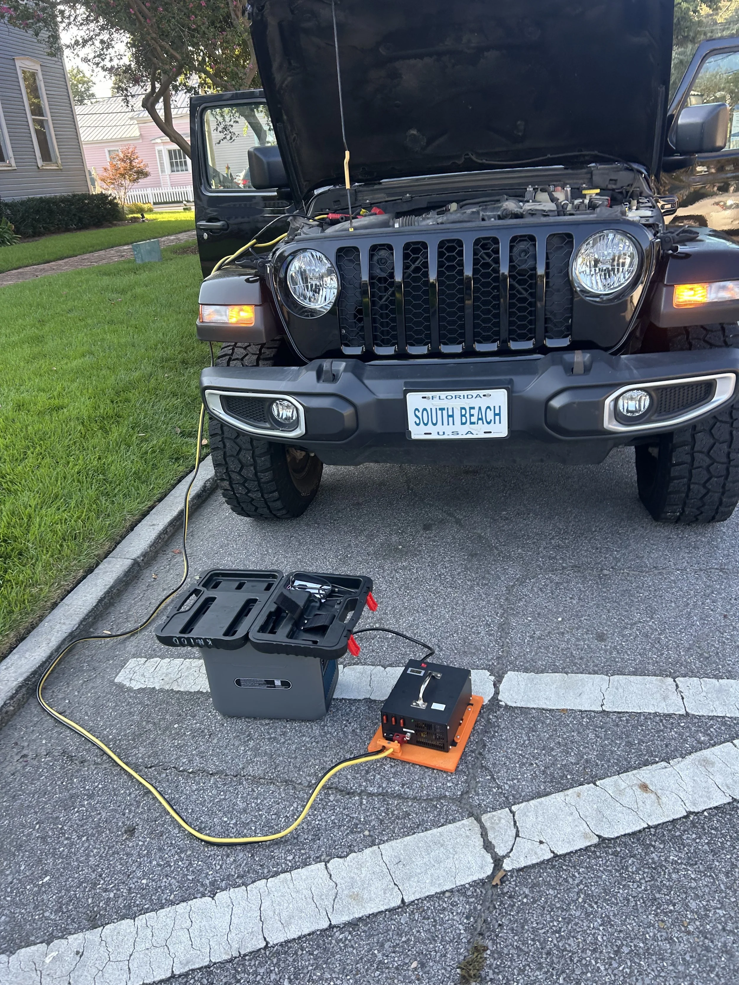 A black Jeep with its hood open, parked in a residential area with houses and trees in the background. There is a diagnostic or repair equipment setup in front of the vehicle on the parking All keys lost. Downtown Pensacola. 2023 Jeep Gladiator.