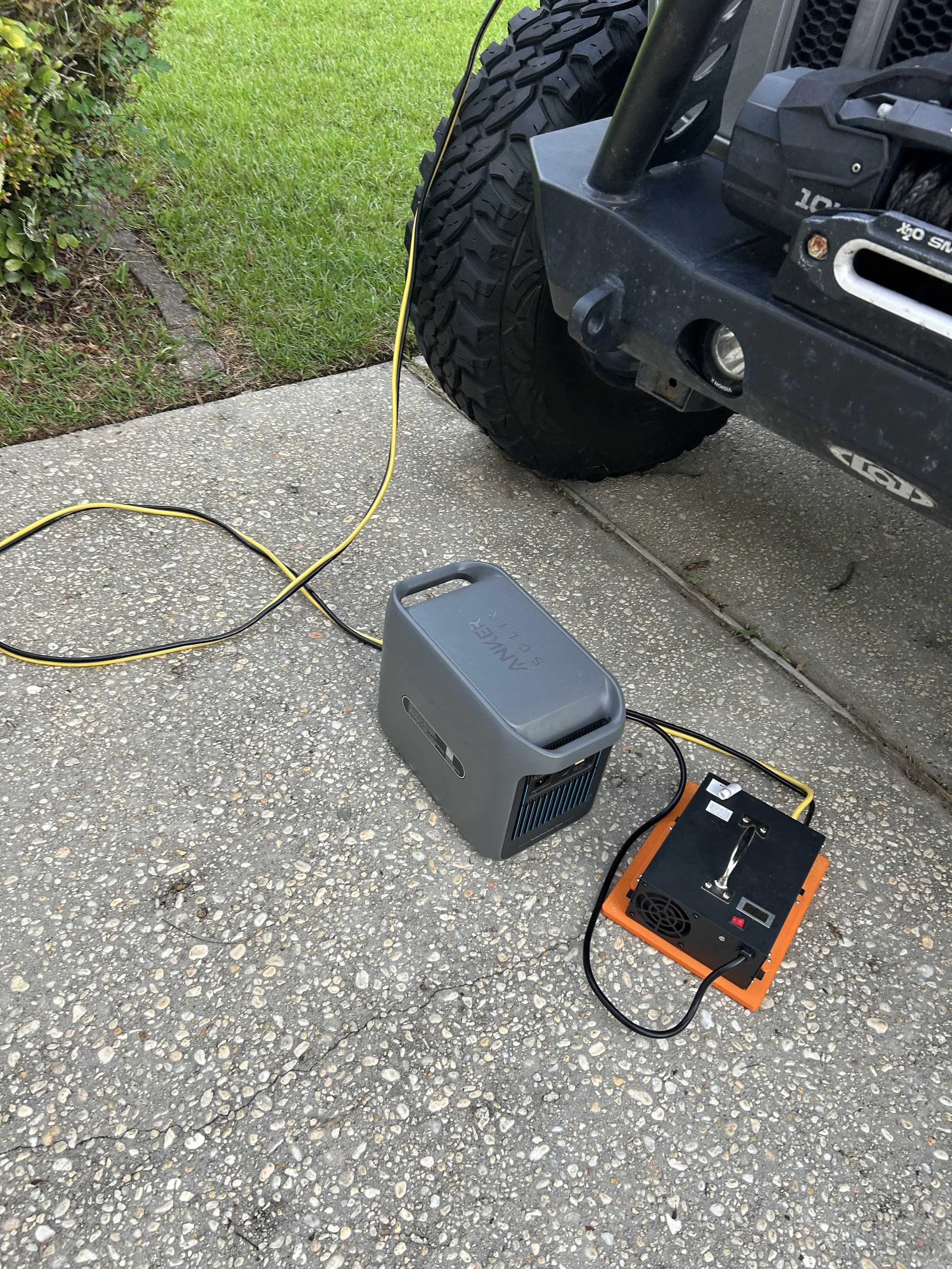 A tire changer machine set up outside on a concrete driveway, connected to a vehicle with a large off-road tire. Various equipment and tools are nearby.