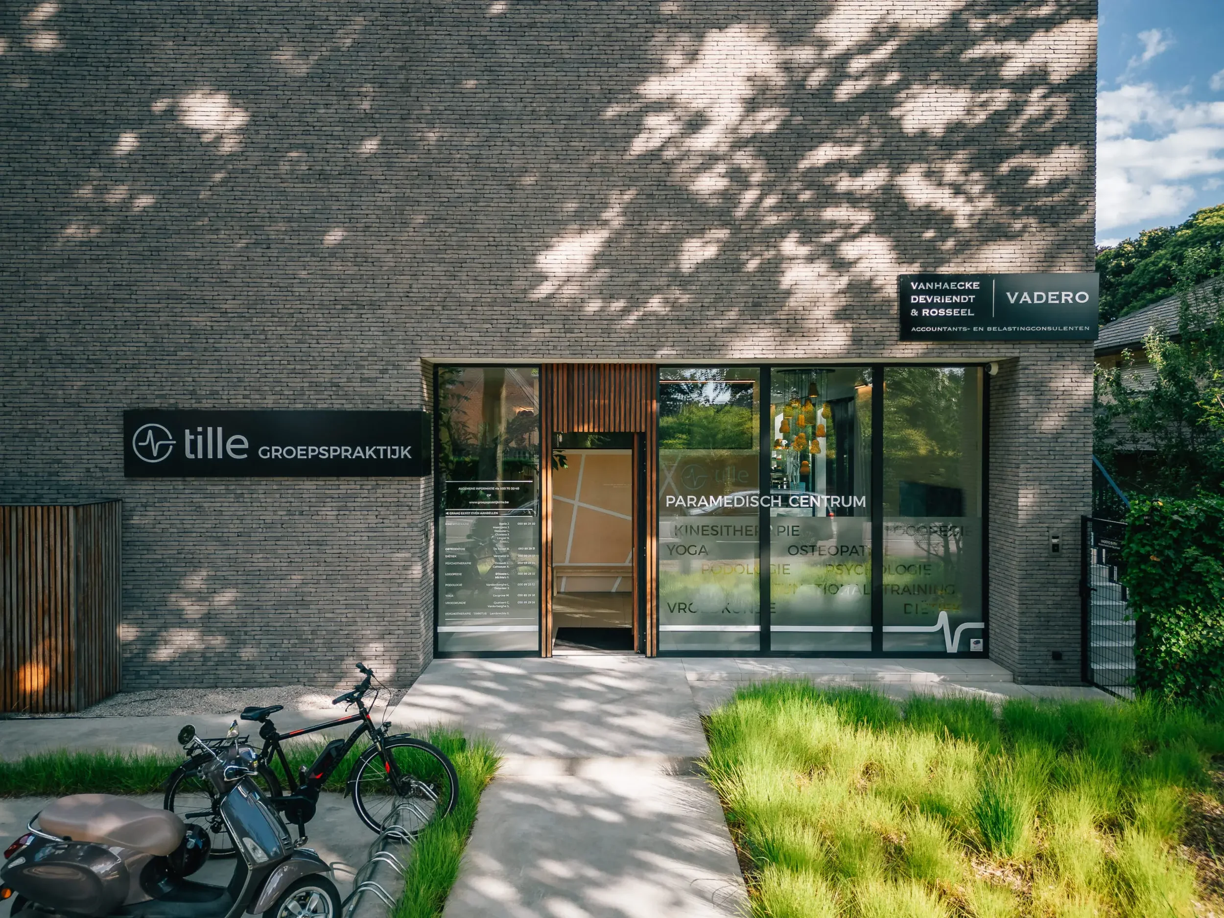 De ingang van een paramedische praktijk met fietsen voor de deur, zicht op een modern gebouw met grote ramen en groene planten rondom. De ramen hebben teksten over paramedisch centrum, osteopathie, yoga en psychologie.