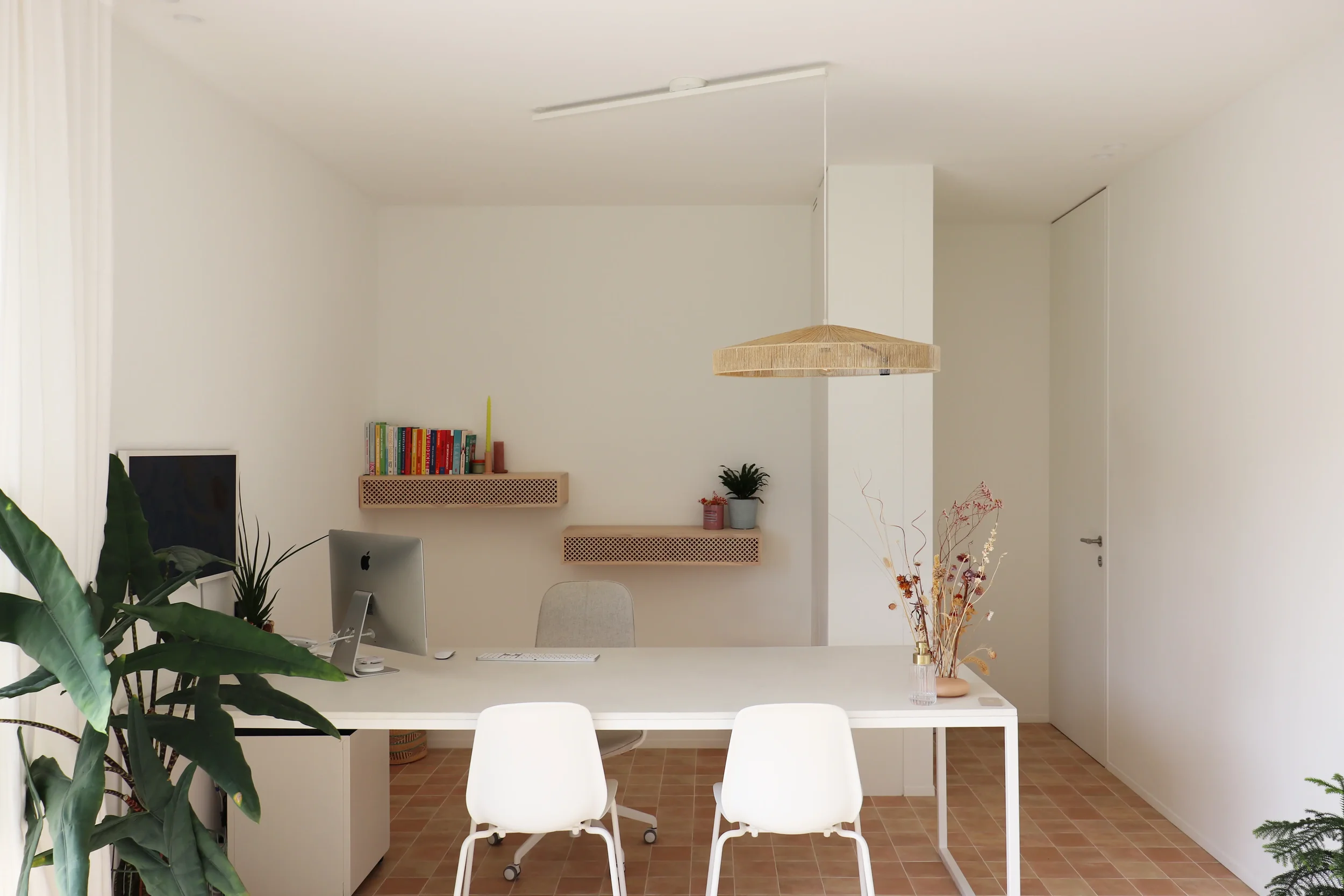 Minimalistisch kantoor met wit bureau, twee witte stoelen, bureaustoel, planten, boeken, en een hanglamp. De vloer bestaat uit terracotta tegels.