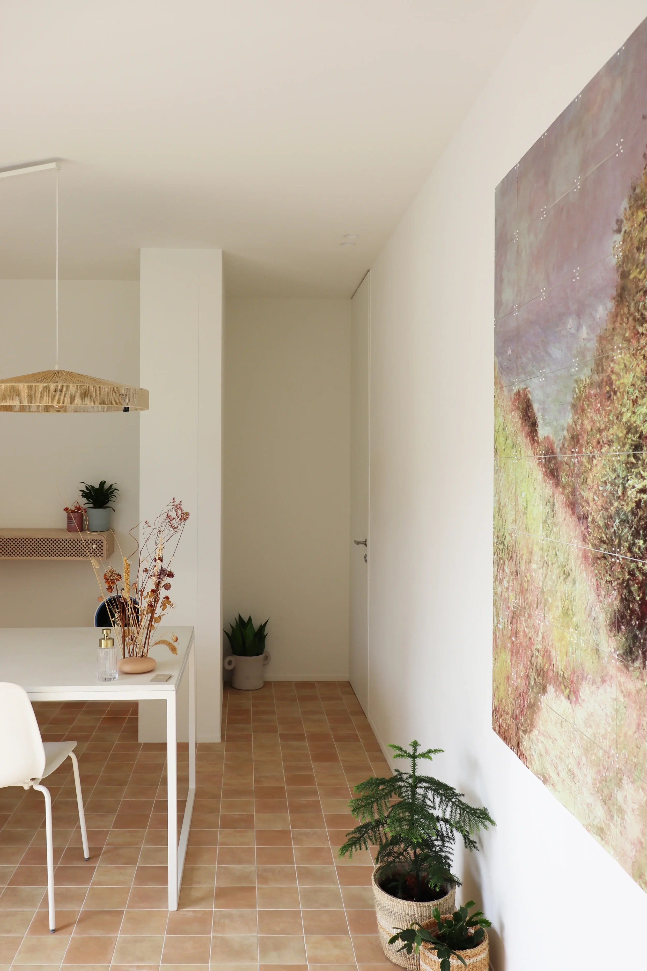 Interieur van een minimalistische, lichte kamer met beige betegelde vloer, witte muren en een houten tafel met vazen en planten, grote muurkunst, en verschillende groene planten in potten.