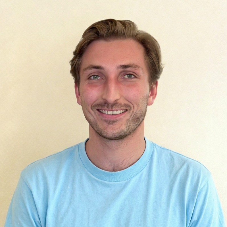 Portret van een lachende man met kastanjebruin haar, blauwe ogen, lichte huid, in een lichte blauwe T-shirt, staand voor een witte muur.