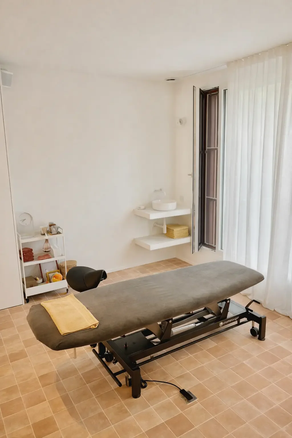 Een behandelingsruimte met een massagebank of tafel voor manuele therapie (in dit geval osteopathie), een kleine ronde lavabo en open ramen met witte gordijnen.