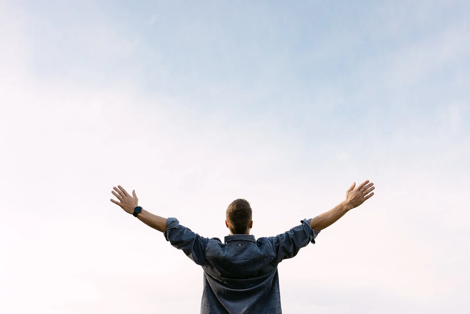 Man met uitgestrekte armen aan de hemel, achtergrond van een heldere, lichte lucht.