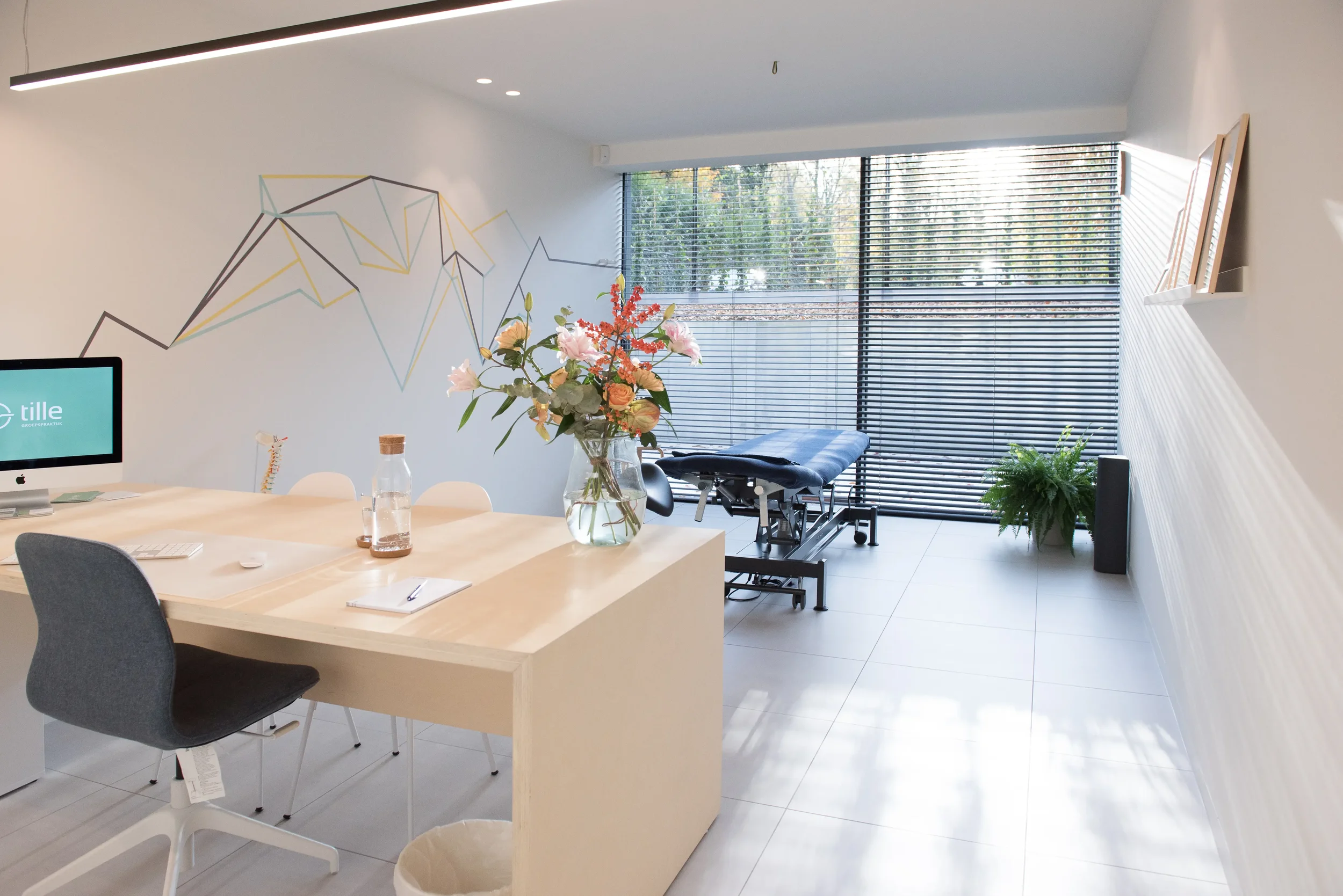 Een behandelkamer met een behandelbank voor manuele therapie/osteopathie, een grote tafel met bloemen in een vaas, een computer en documentaire materialen, grote ramen met horizontale jaloezieën, een groen plant aan de muur, en een wandtekening