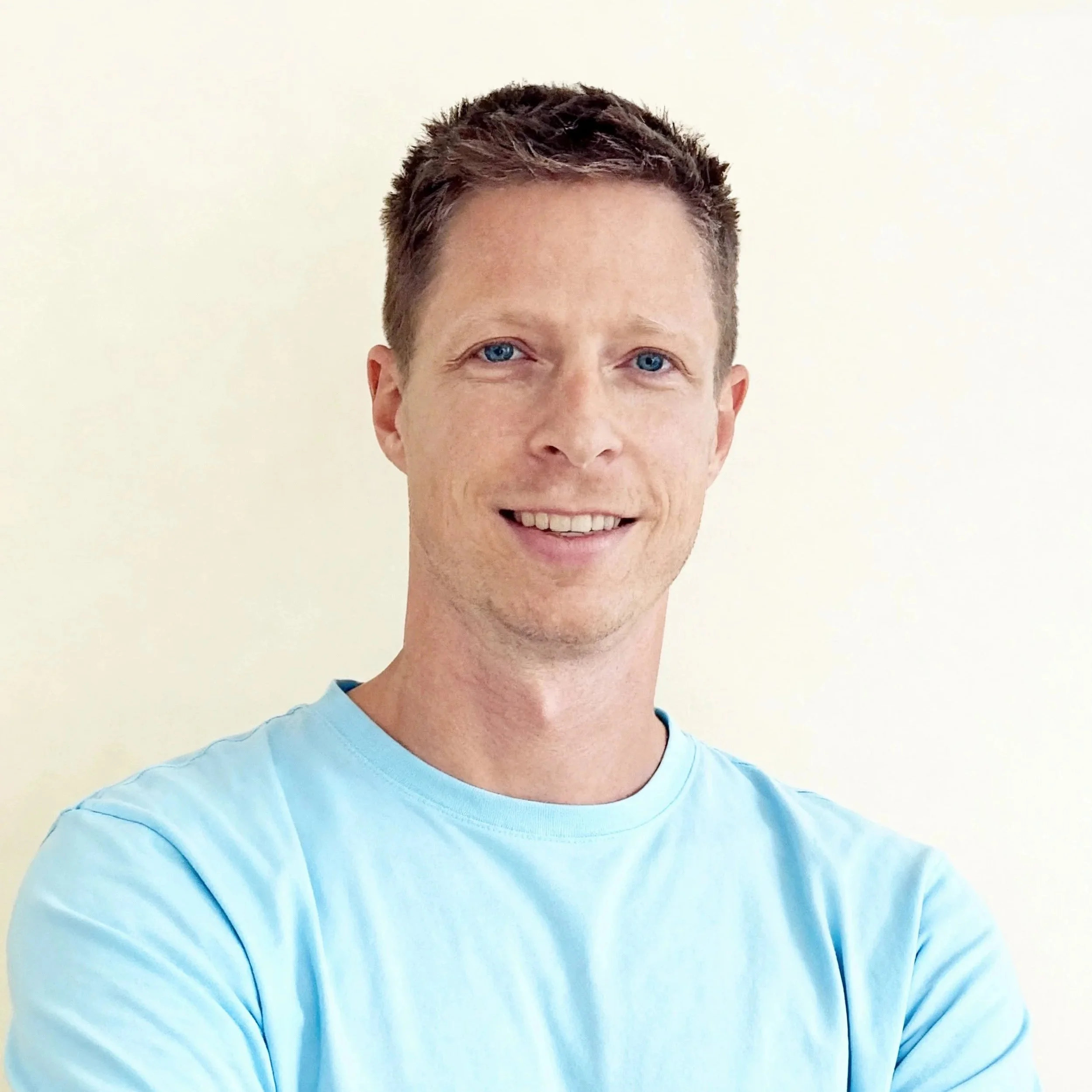 Portret van een man met blauwe ogen en kort bruin haar, lachend, gekleed in een lichtblauw T-shirt, staand tegen een lichte achtergrond.