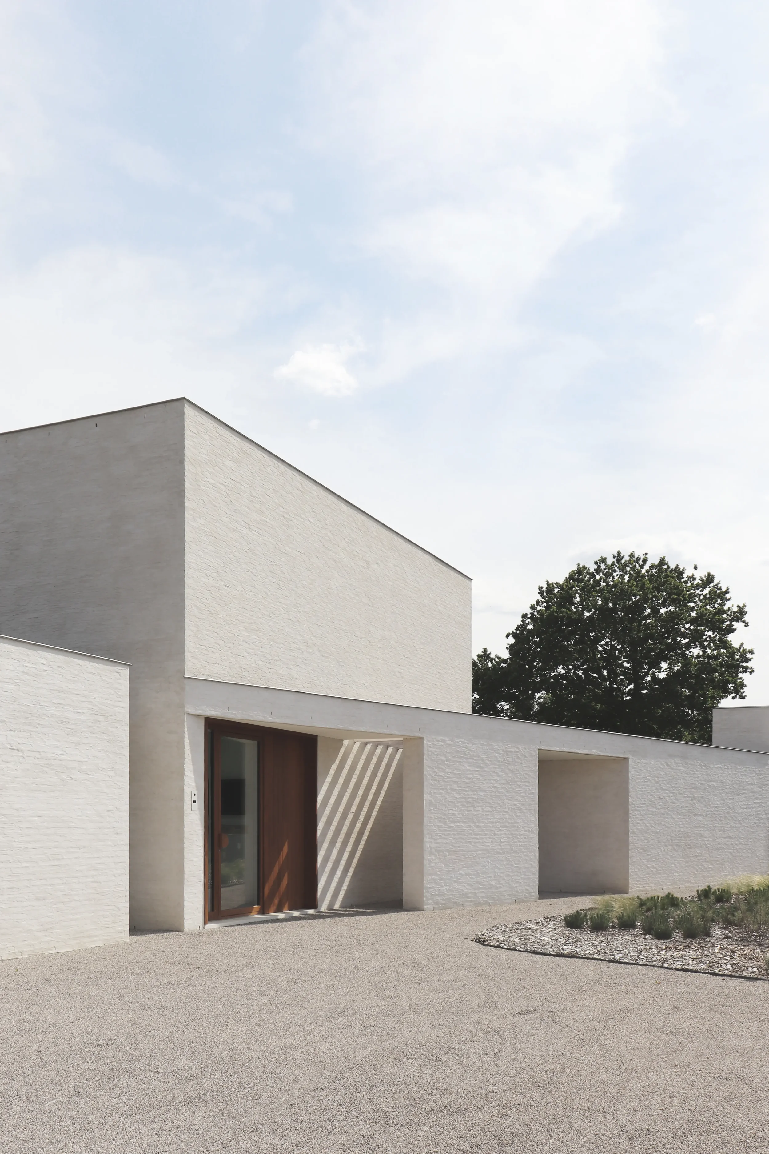 Moderne witte woning met een houtkleurige deur en een tuin met gras en planten, onder een lichte bewolkte hemel.