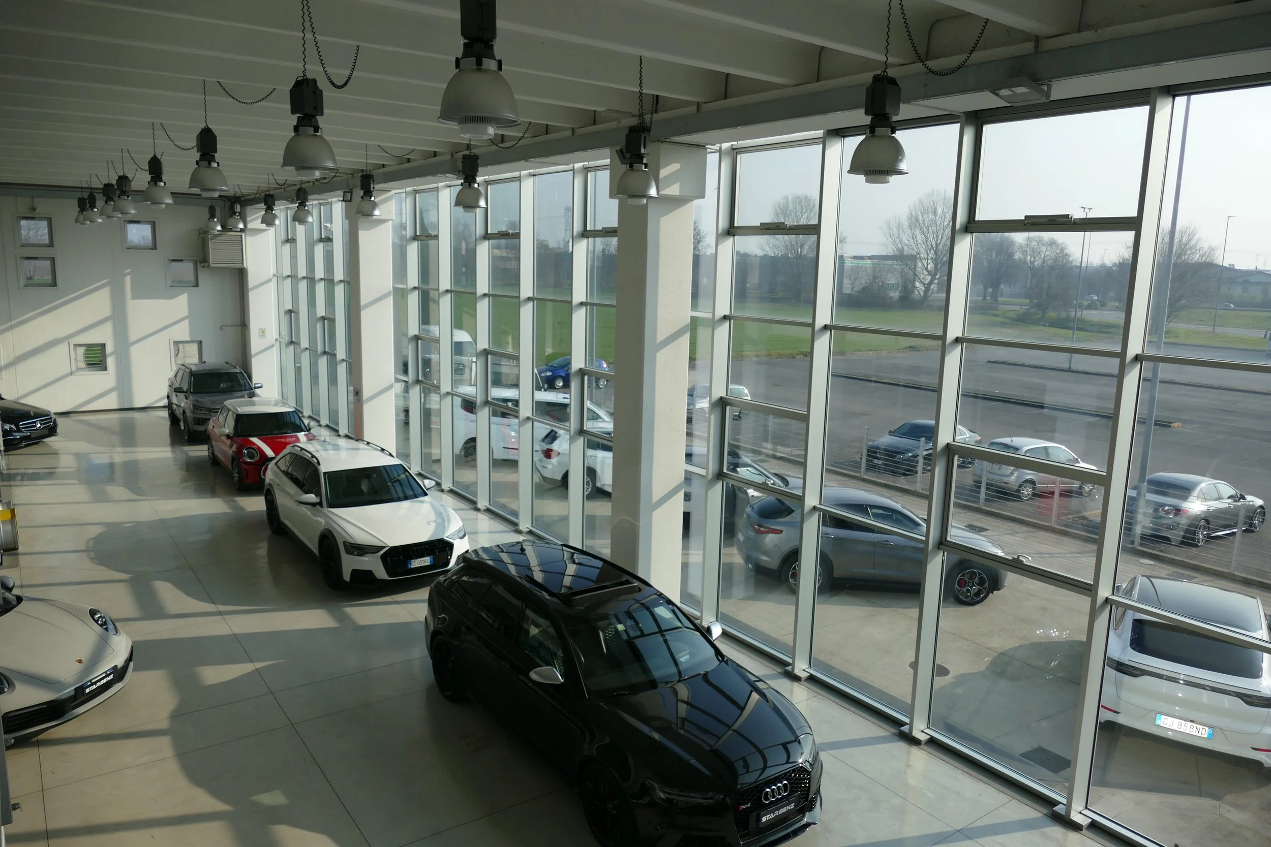 Interno di un'autoagenzia con diversi veicoli esposti, tra cui un Audi nero, una Porsche beige e altre auto di varie marche e colori, con grandi finestre e luci al soffitto.