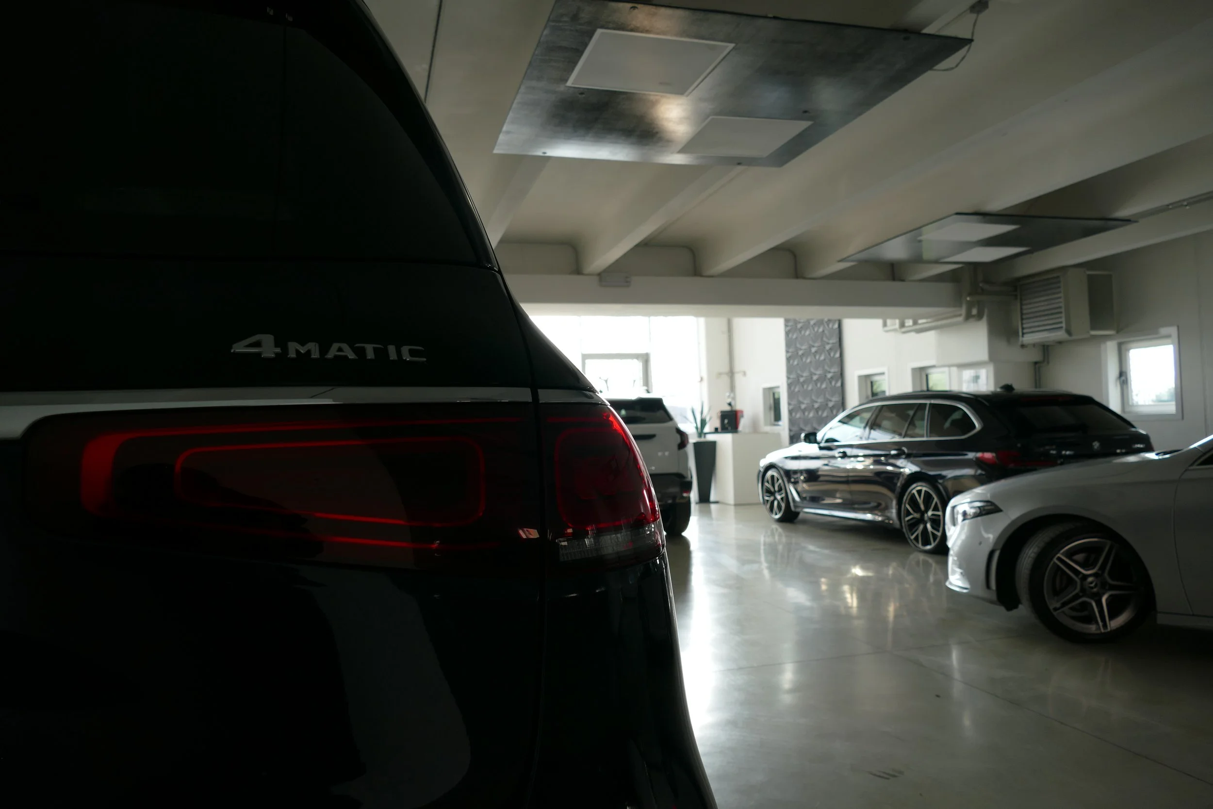 Auto di lusso in un garage, con un veicolo nero sul lato sinistro e altre tre auto di colore chiaro sul lato destro, illuminato dalla luce naturale attraverso le finestre.