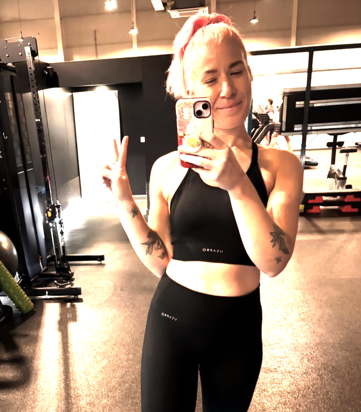 A woman with pink hair wears gym gear. She has pink hair. She is in a gym in folkestone. She makes a peace gesture with her hands