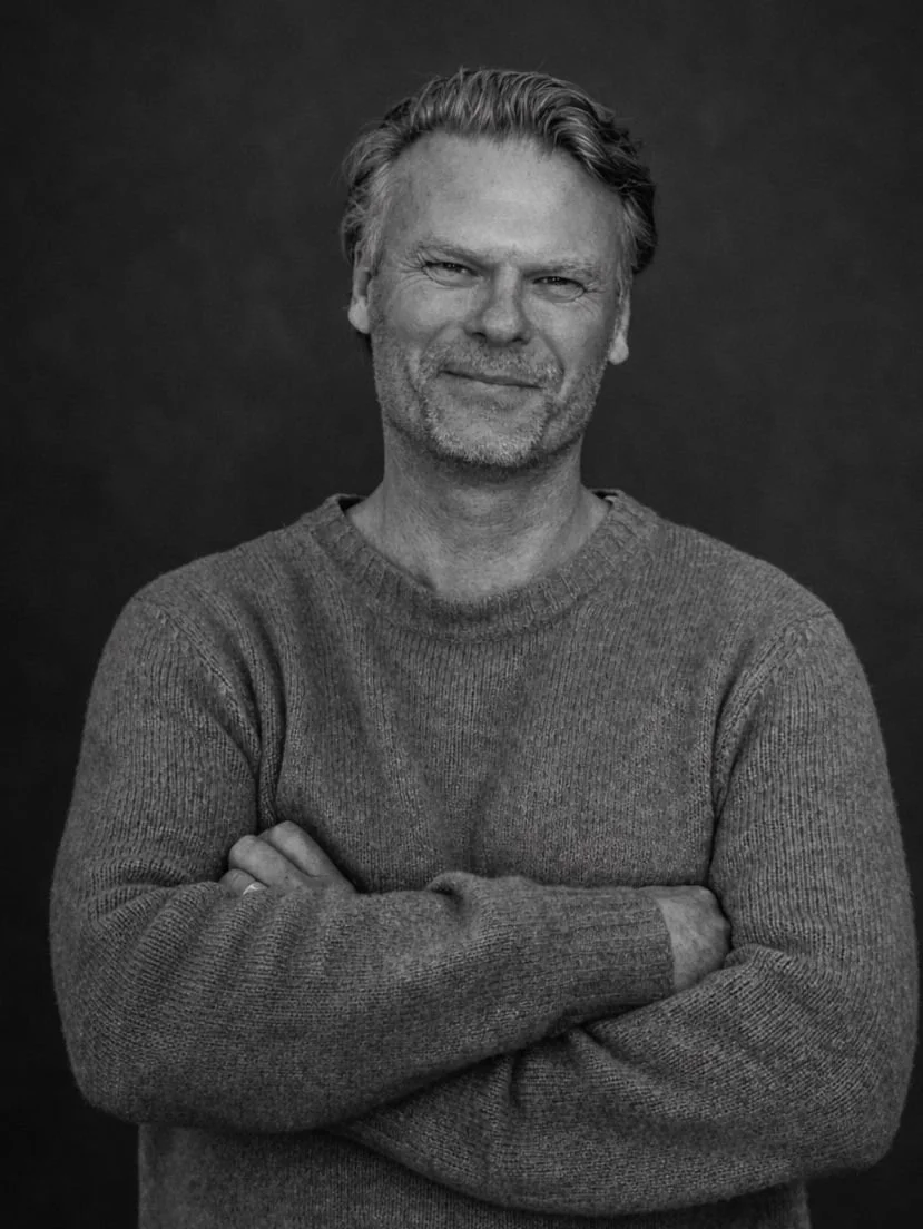 Black and white portrait of a middle-aged man with light hair, beard, and mustache, wearing a knit sweater, smiling with arms crossed, against a dark background.