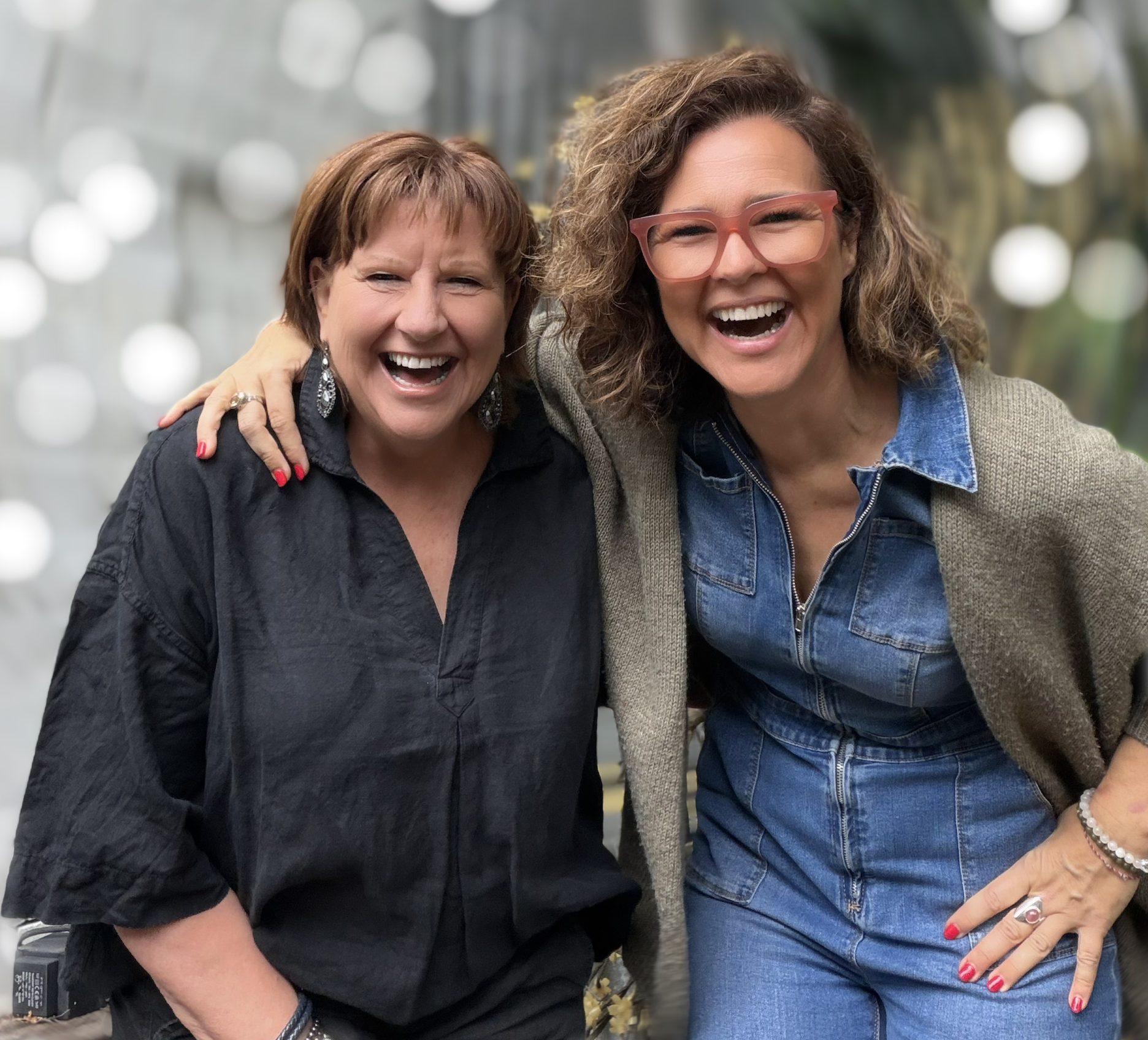 Two women with short brown hair, smiling and laughing, hugging each other in an outdoor setting with bokeh lights in the background. El Chambers and Nicole Perry offering women's spiritual retreats and empowerment coaching in Victoria.