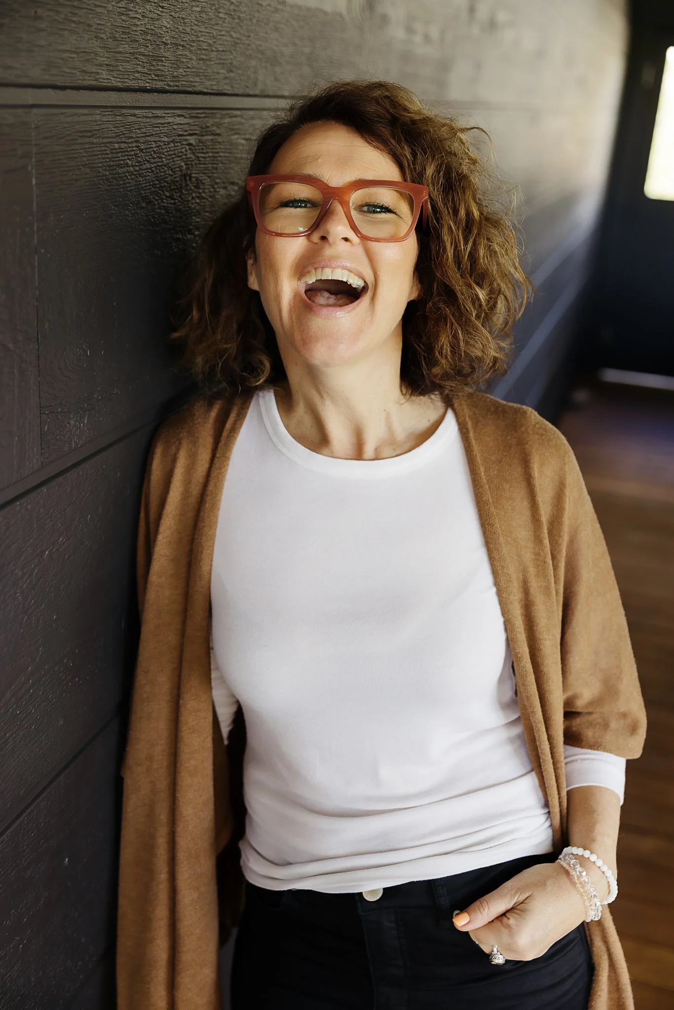 A woman with curly brown hair wearing glasses, a white shirt, and a brown cardigan, laughing and leaning against a dark wood wall inside a room. El Chambers and Nicole Perry offering women's spiritual retreats and empowerment coaching in Victoria.