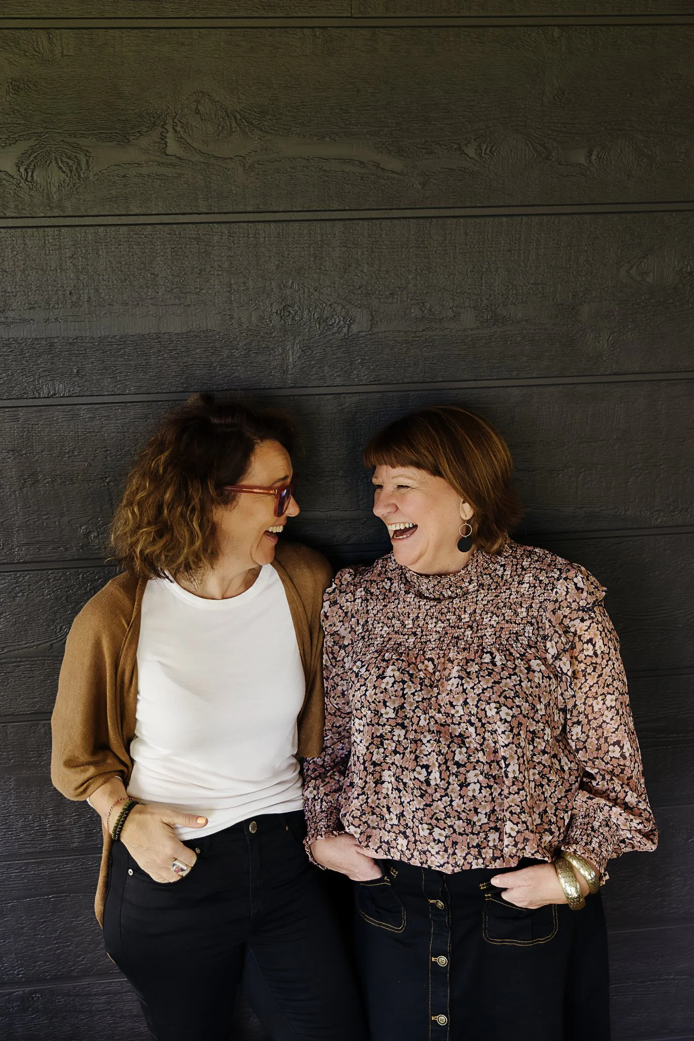 Two women standing close and smiling at each other in front of a black wooden wall.  El Chambers and Nicole Perry offering women's spiritual retreats and empowerment coaching in Victoria.