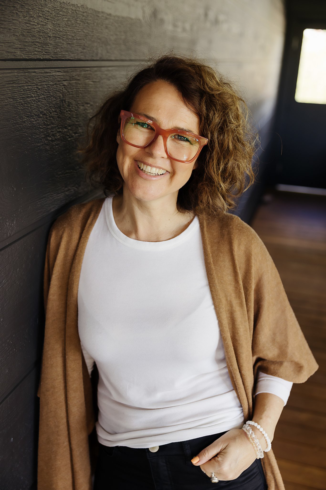 A woman with curly brown hair and glasses smiling, wearing a white shirt, black jeans, standing against a dark wall. El Chambers and Nicole Perry offering women's spiritual retreats and empowerment coaching in Victoria