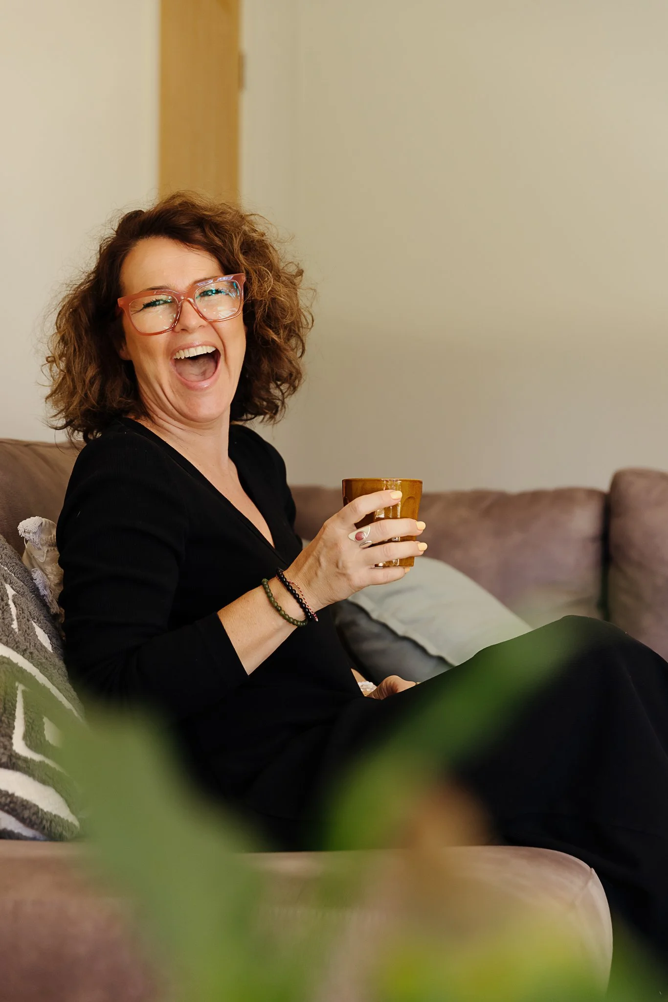 A woman with curly brown hair is laughing and holding a small cup while sitting on a sofa, in a room with beige walls. El Chambers and Nicole Perry offering women's spiritual retreats and empowerment coaching in Victoria.