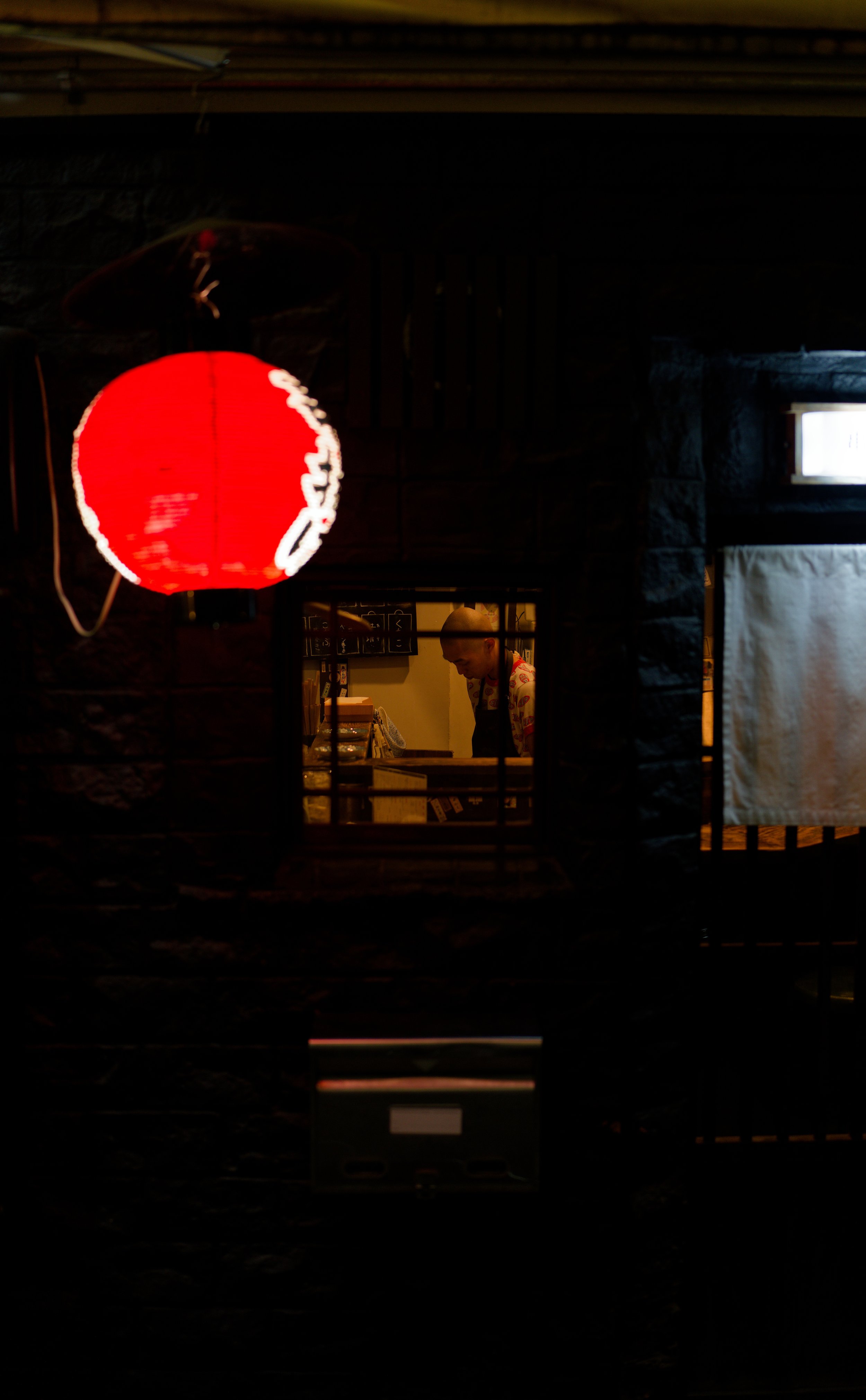 View of a person inside a small room, seen through a window with a grid, with a red lantern hanging outside.
