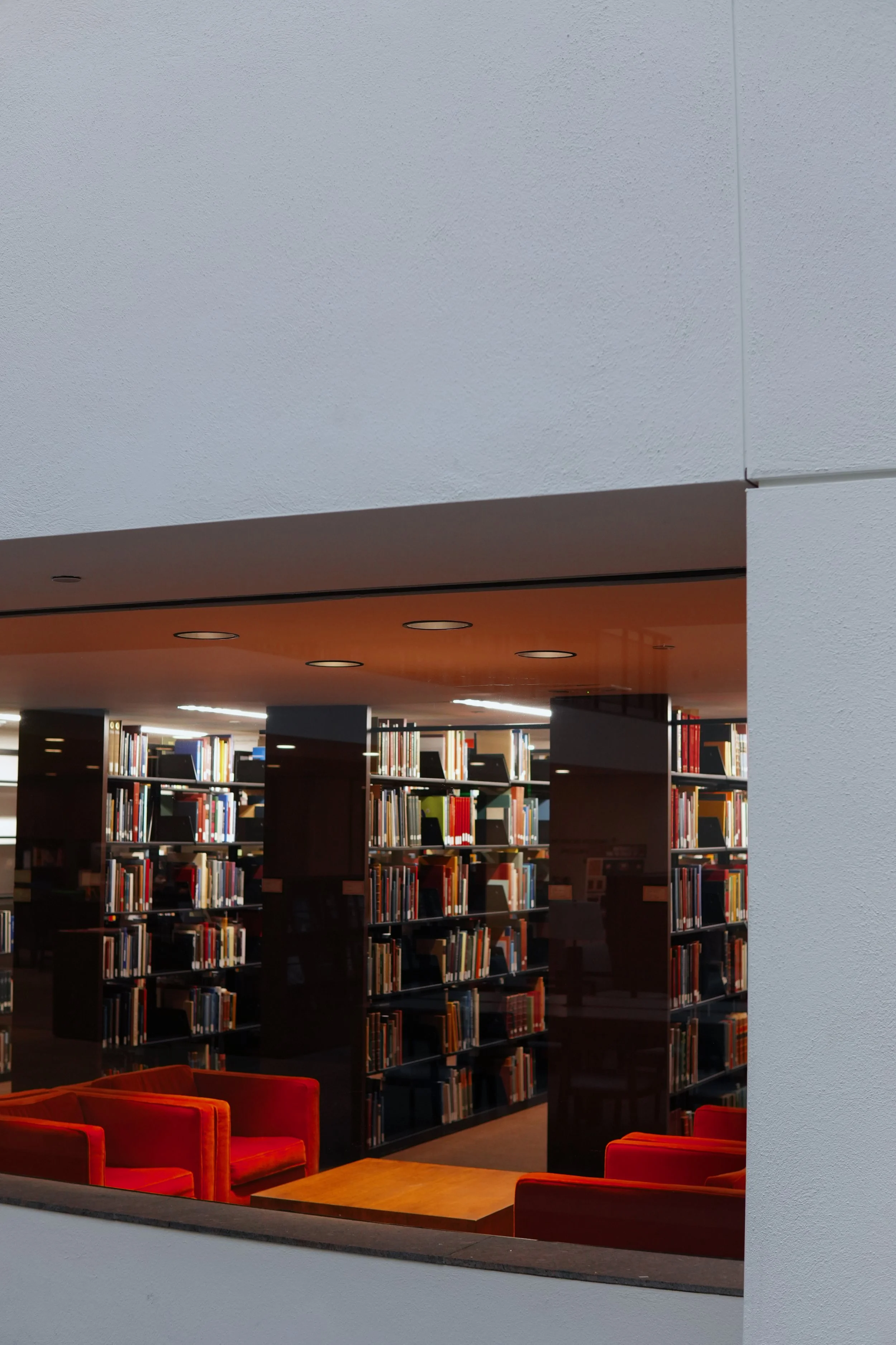 Interior view of a library with bookshelves, orange armchairs, and a table seen through a large rectangular window.