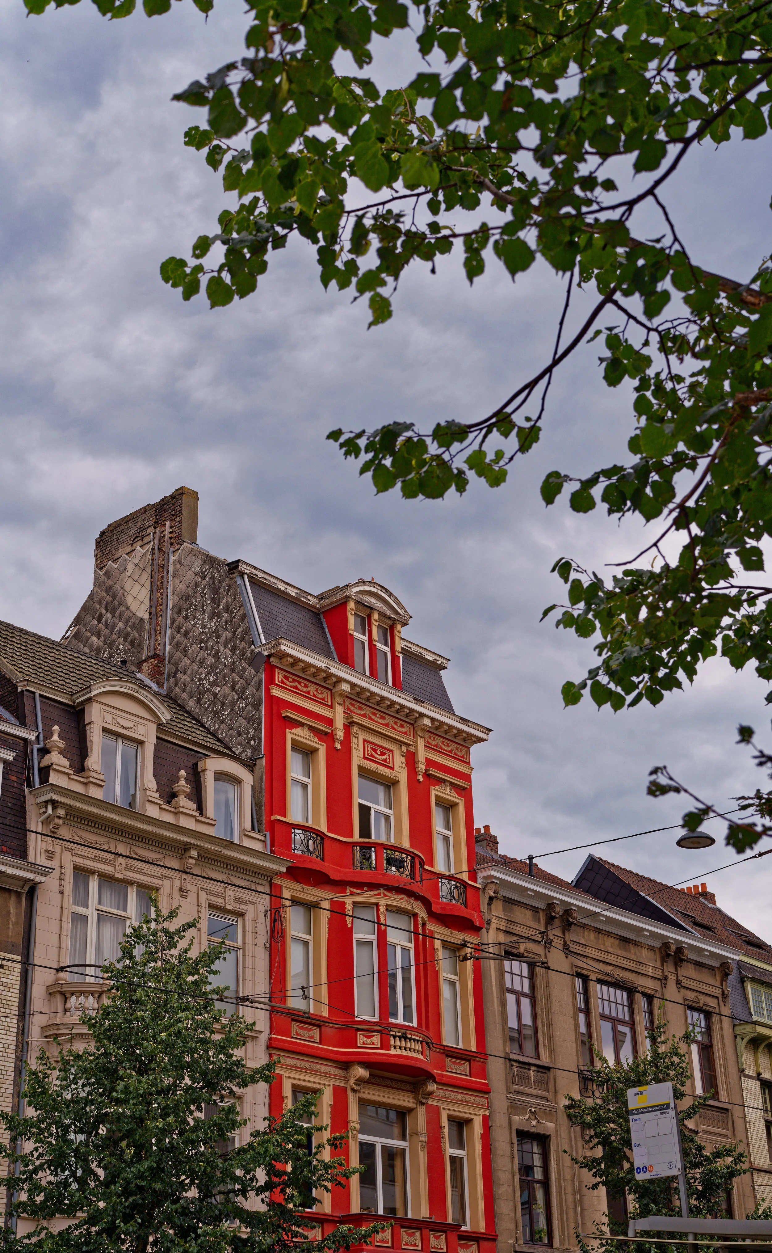 Colorful, ornate multi-story building with red and beige details, situated between other historic buildings, under a cloudy sky with greenery in the foreground.