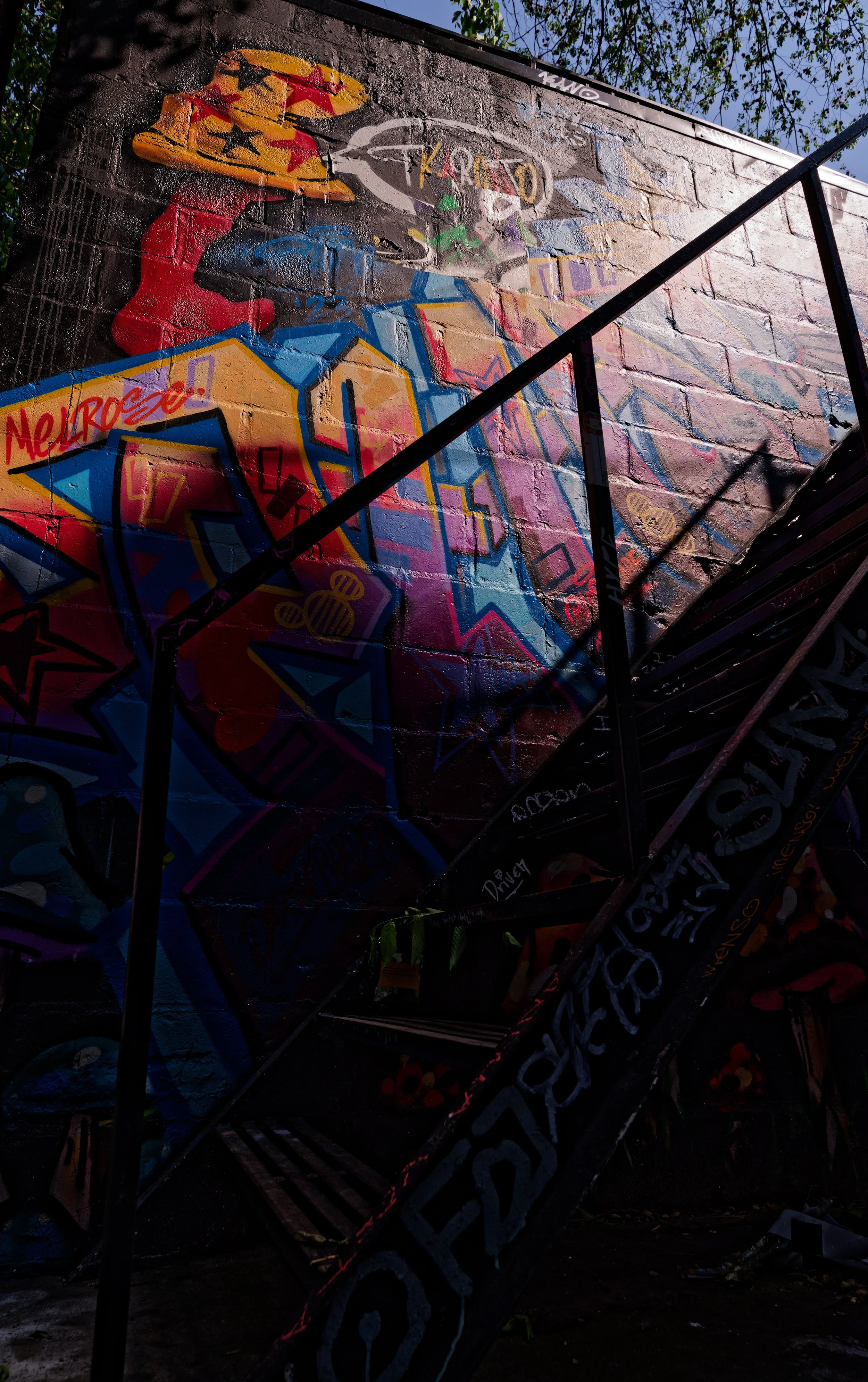 Colorful graffiti art on a brick wall featuring a woman with a hat and the word "Melrose." Visible staircase in front of the wall with graffiti.