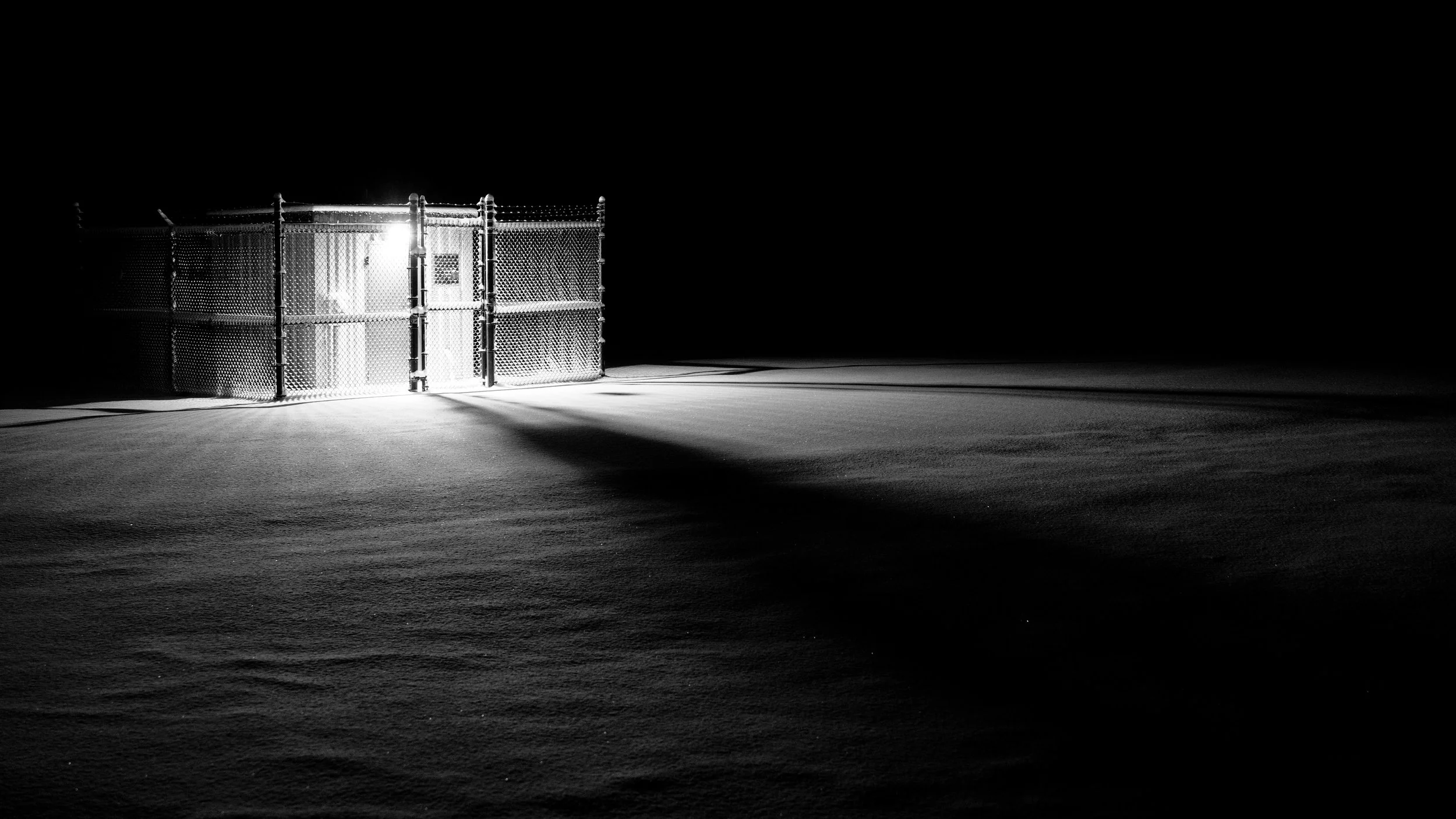 A metallic gate illuminated by a bright light in the darkness, casting long shadows on a snow-covered ground.
