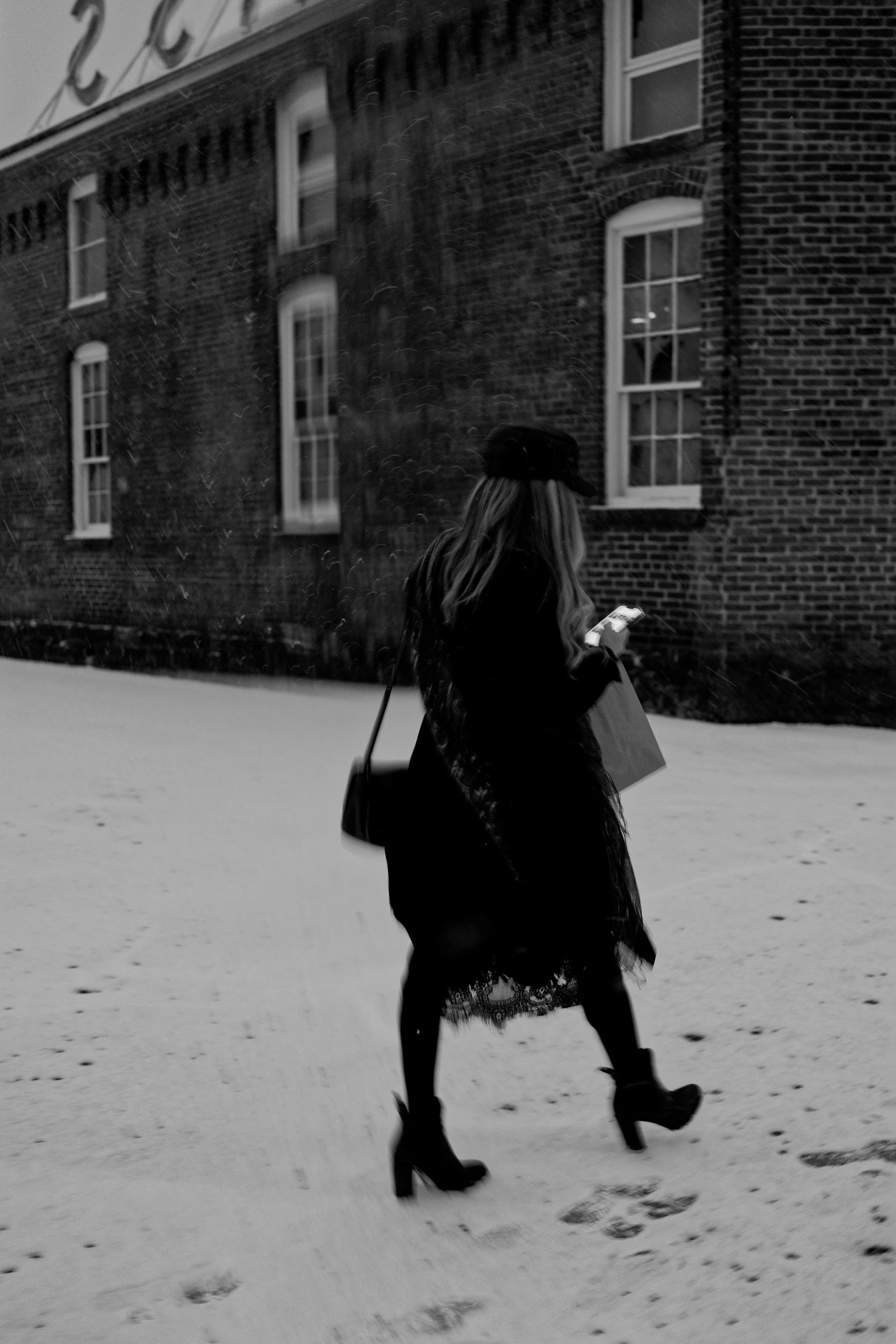 A woman walking on a snow-covered sidewalk, looking at her phone while carrying a shopping bag, wearing high heels, a coat, a hat, and a dress, with a brick building in the background.