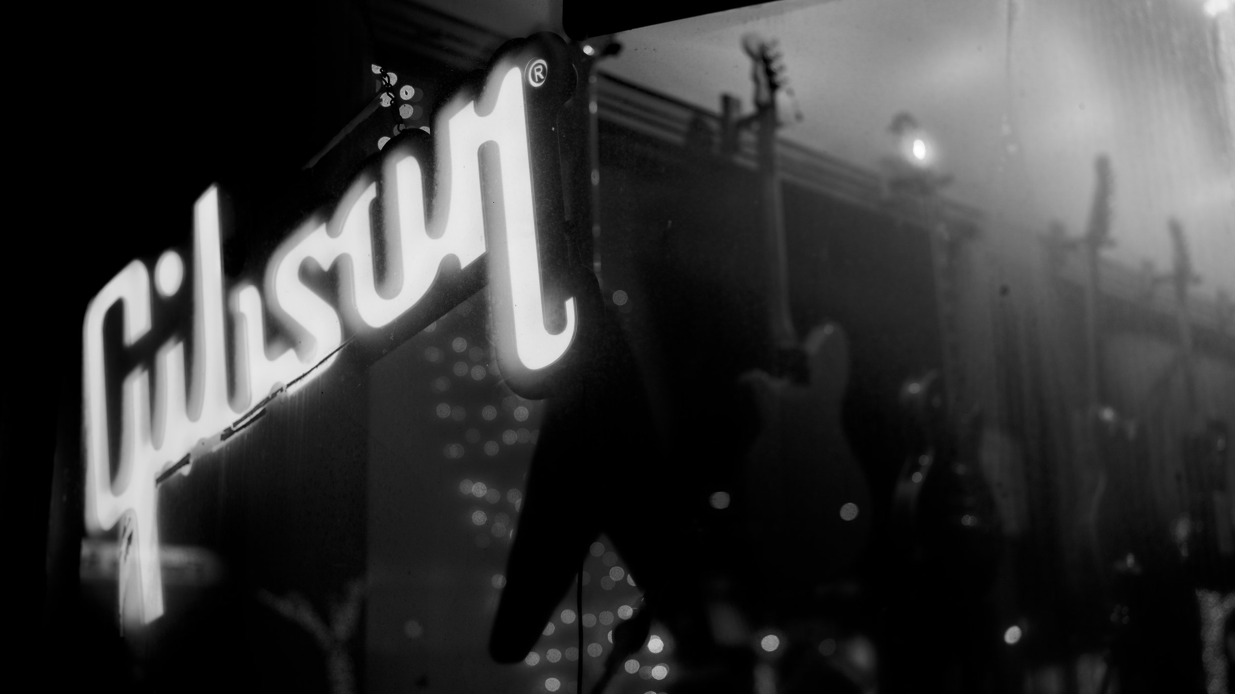 Black and white photo of a lit sign that reads 'Gibson,' with guitars hanging in the background.