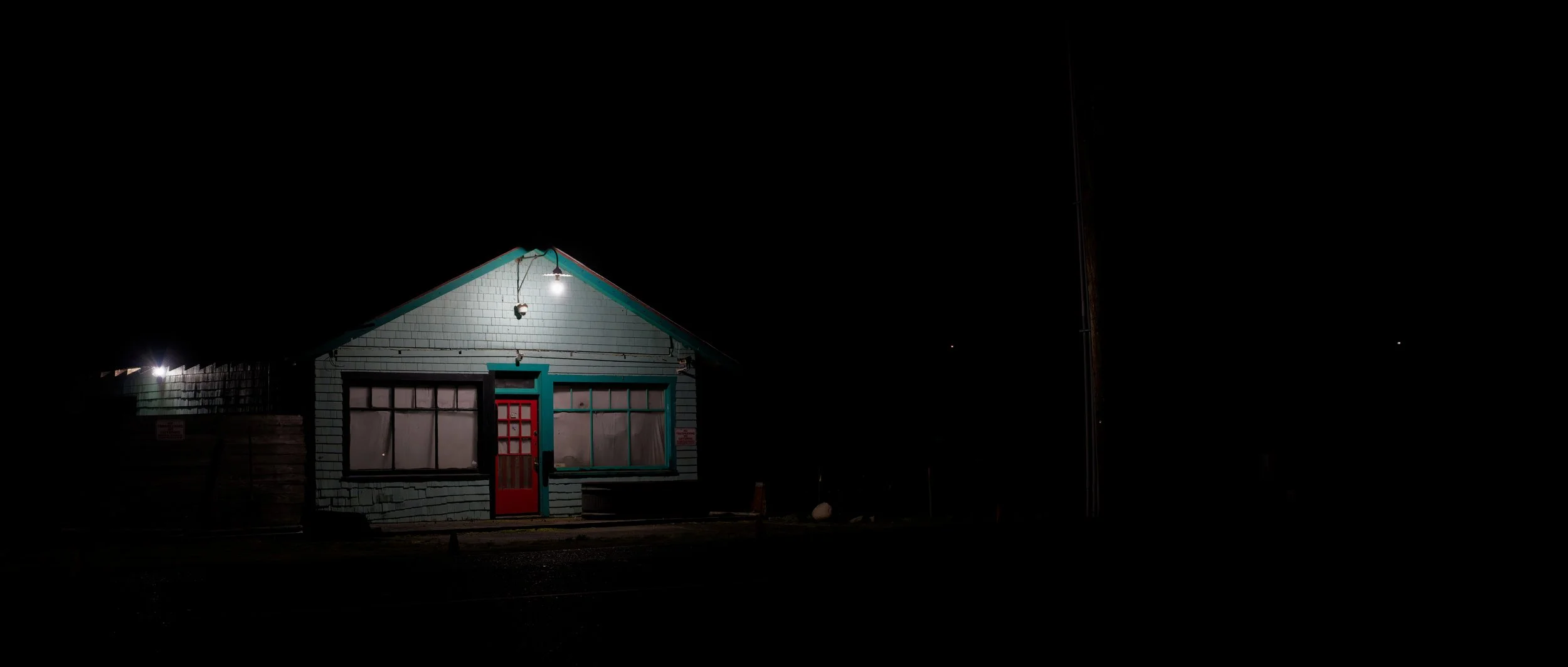 A small house illuminated at night with a red door, large front windows, and a single light hanging outside.