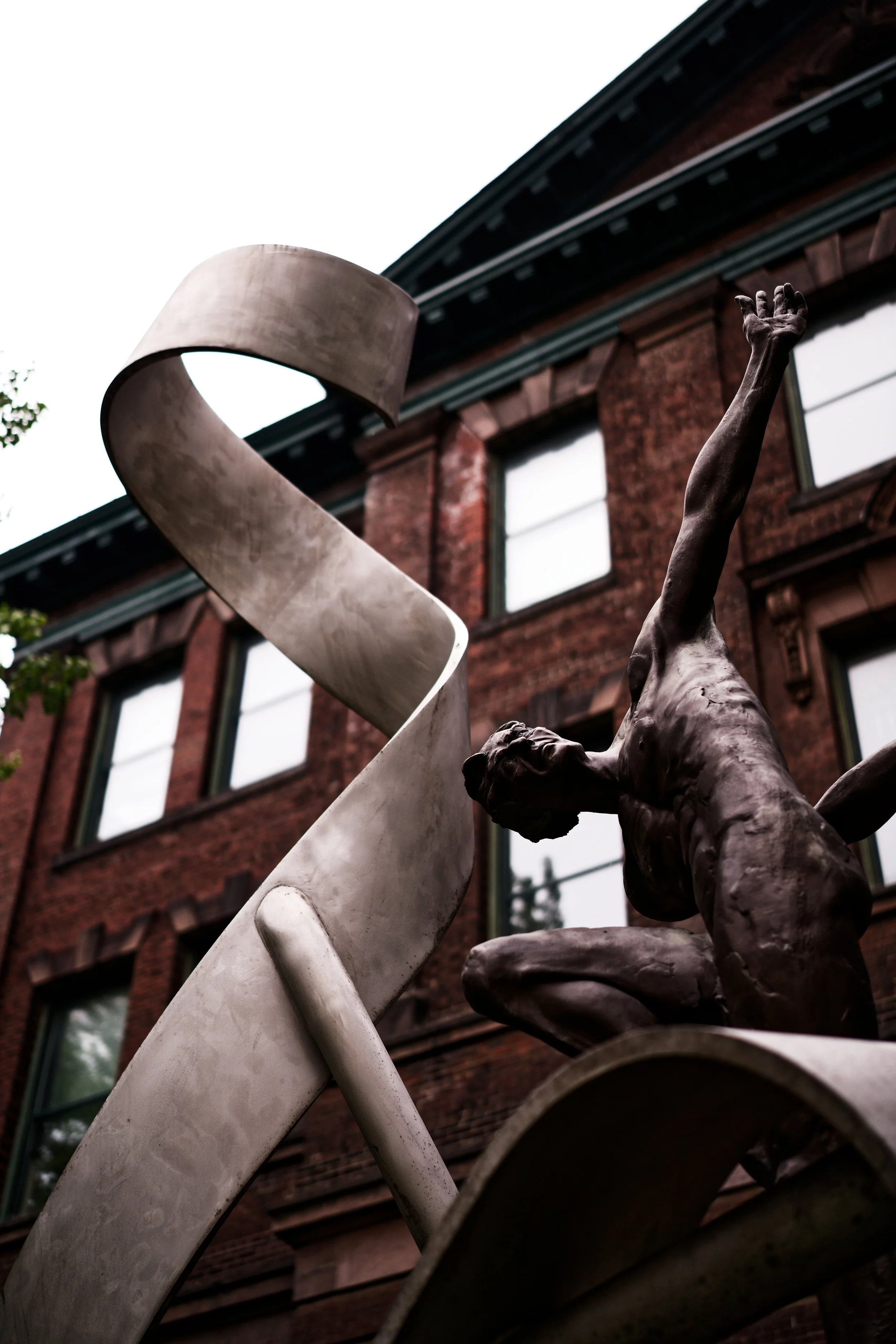A bronze sculpture of a dynamic dancer with outstretched arm, set against a red brick building. A large, twisted metallic ribbon element is part of the sculpture.
