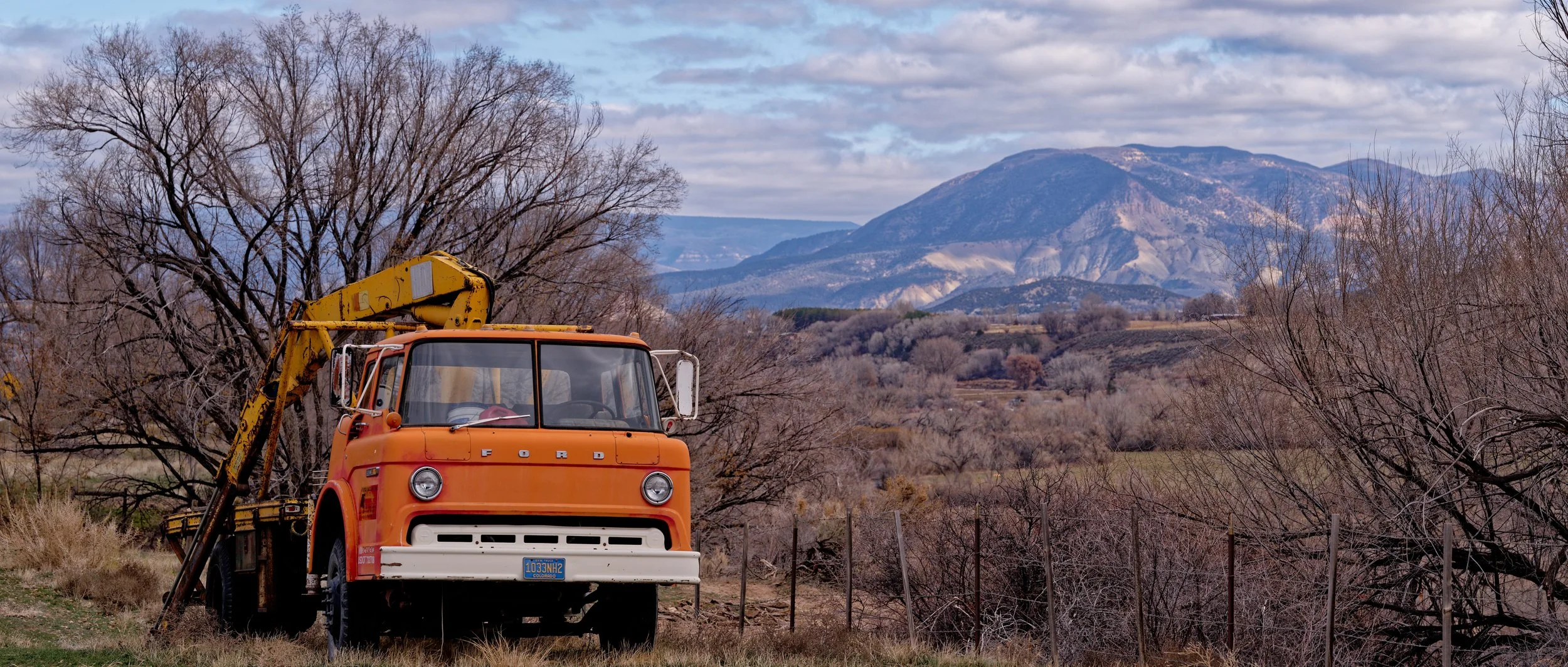 Orange vintage Ford truck with a yellow crane parked in a rural landscape with leafless trees, rolling hills, and mountains in the background under a partly cloudy sky.