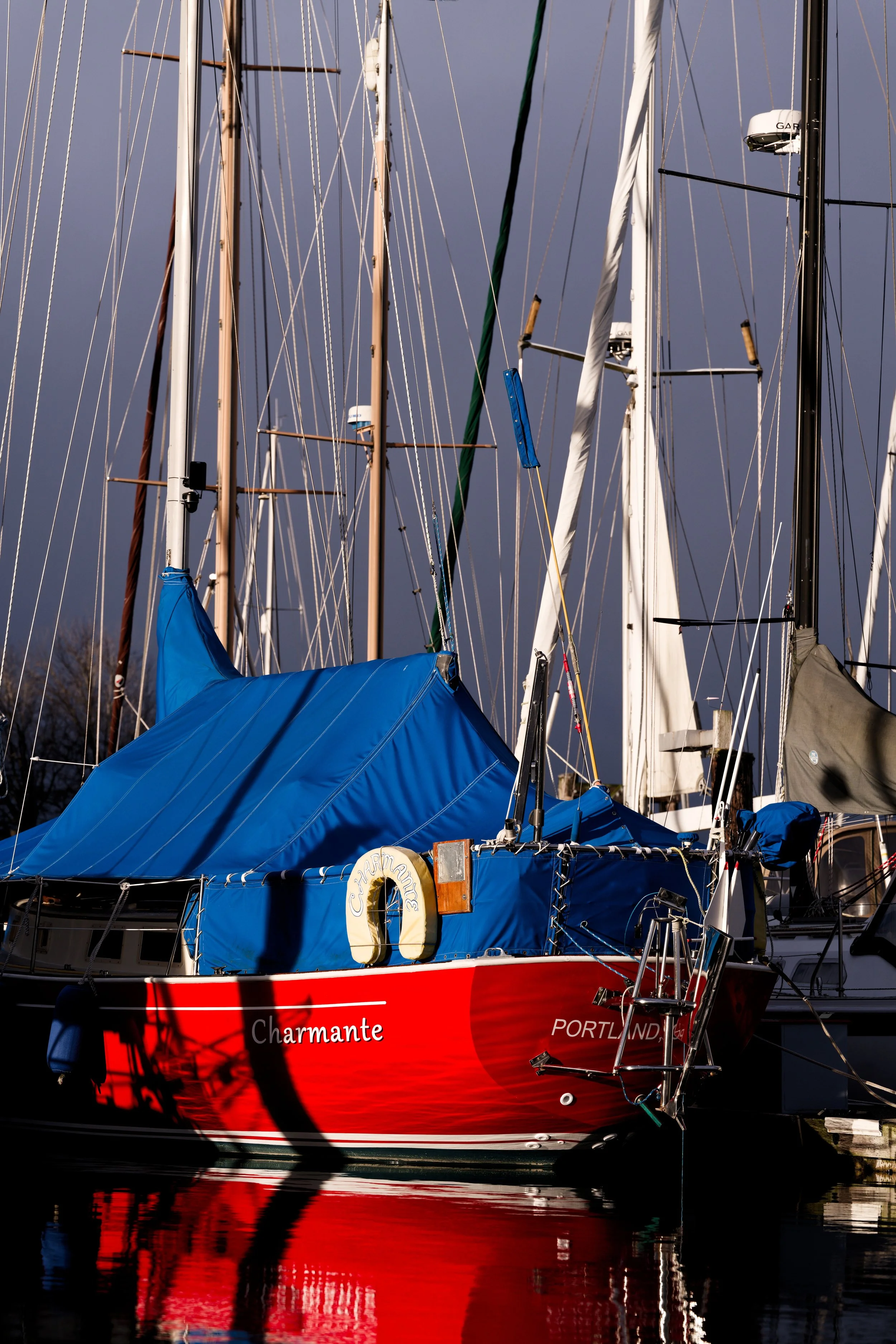 A red sailboat named 'Charmante' docked at a marina with a blue cover on the cabin and multiple masts with rigging and sails. The water reflects the boat and the overcast sky.