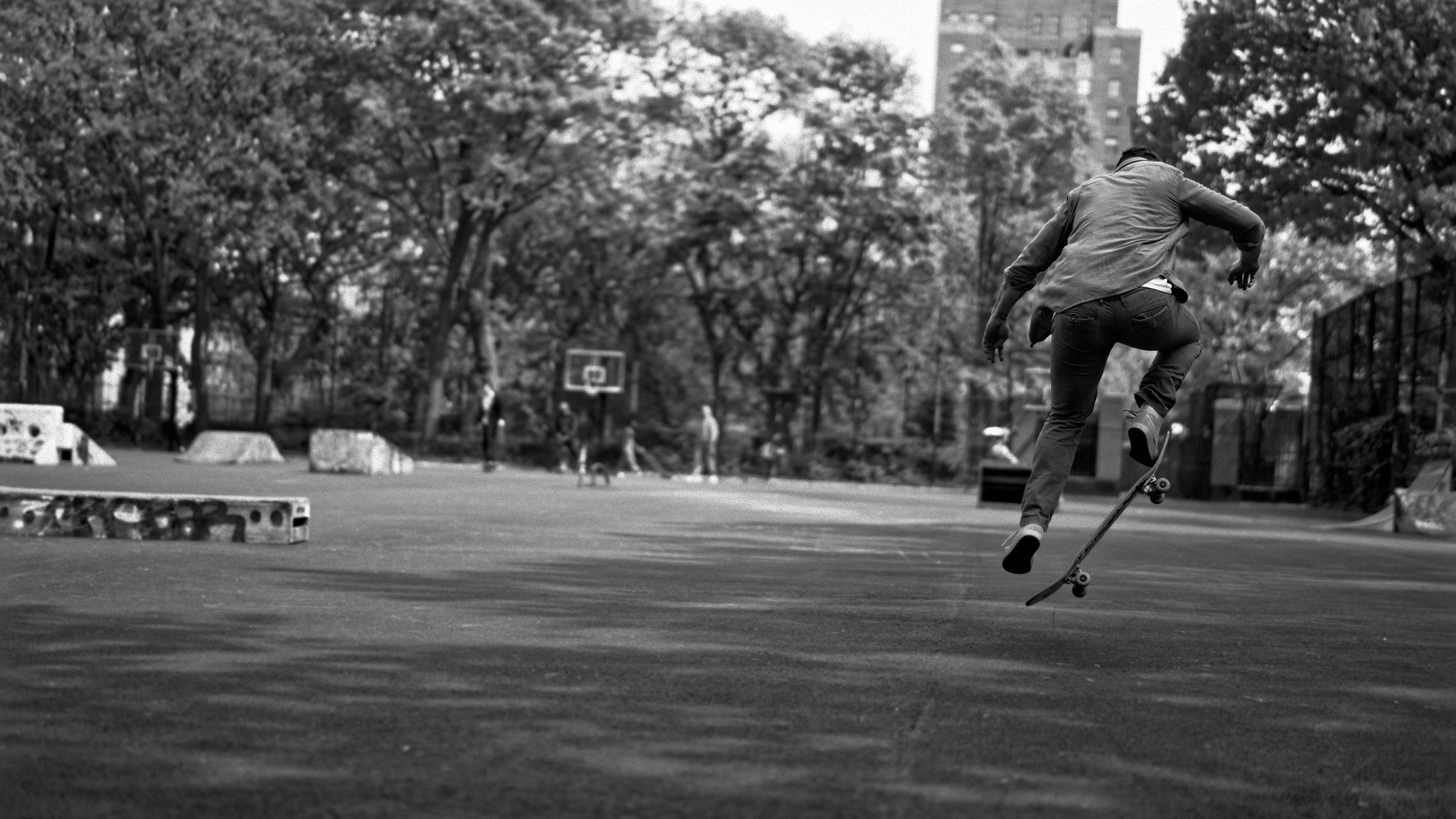 Young skateboarder performing a trick at an outdoor skate park with trees and other skaters in the background