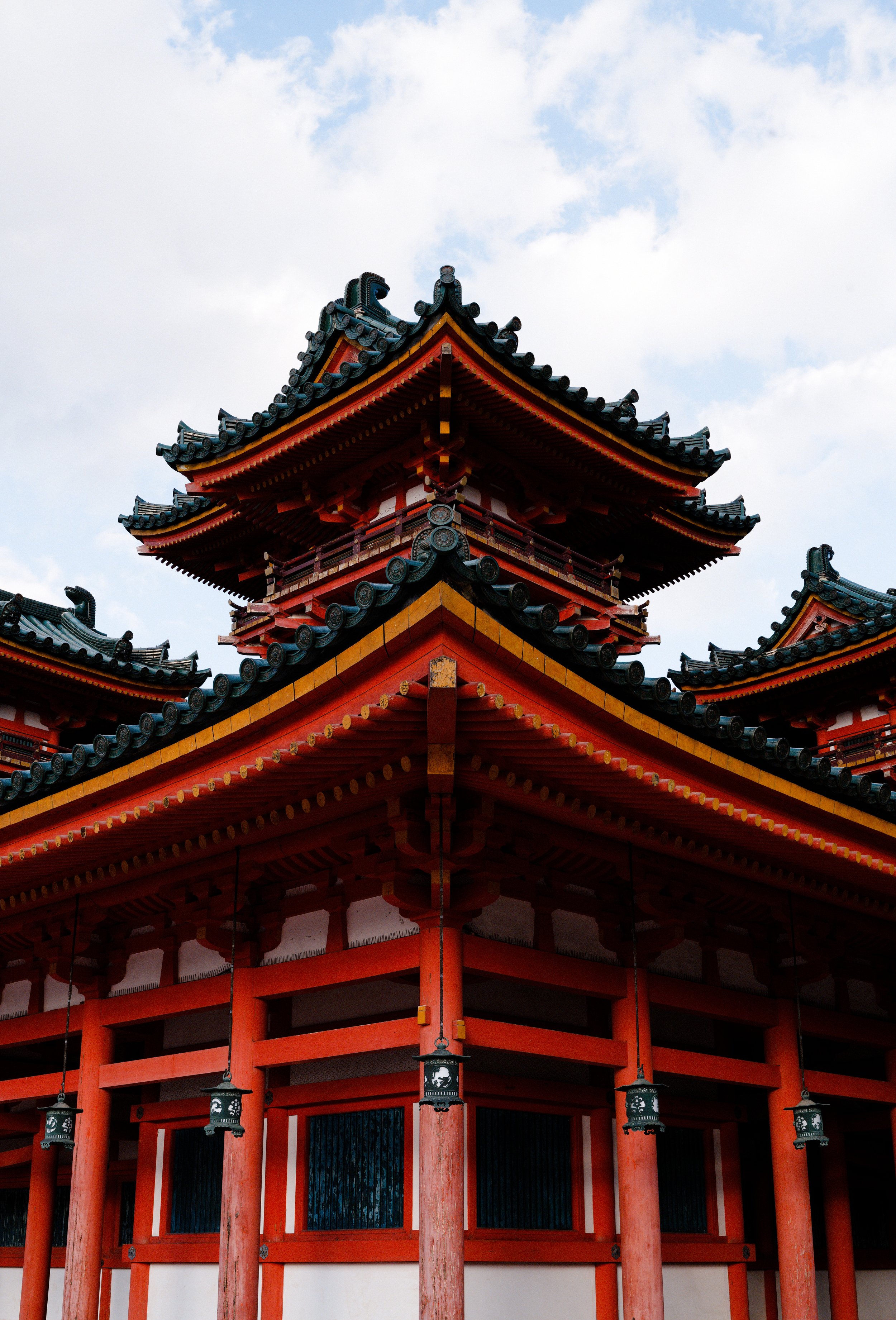 Traditional Japanese temple with red wooden structure, black tiled curved roofing, and paper lanterns hanging from the porch, set against a cloudy sky.