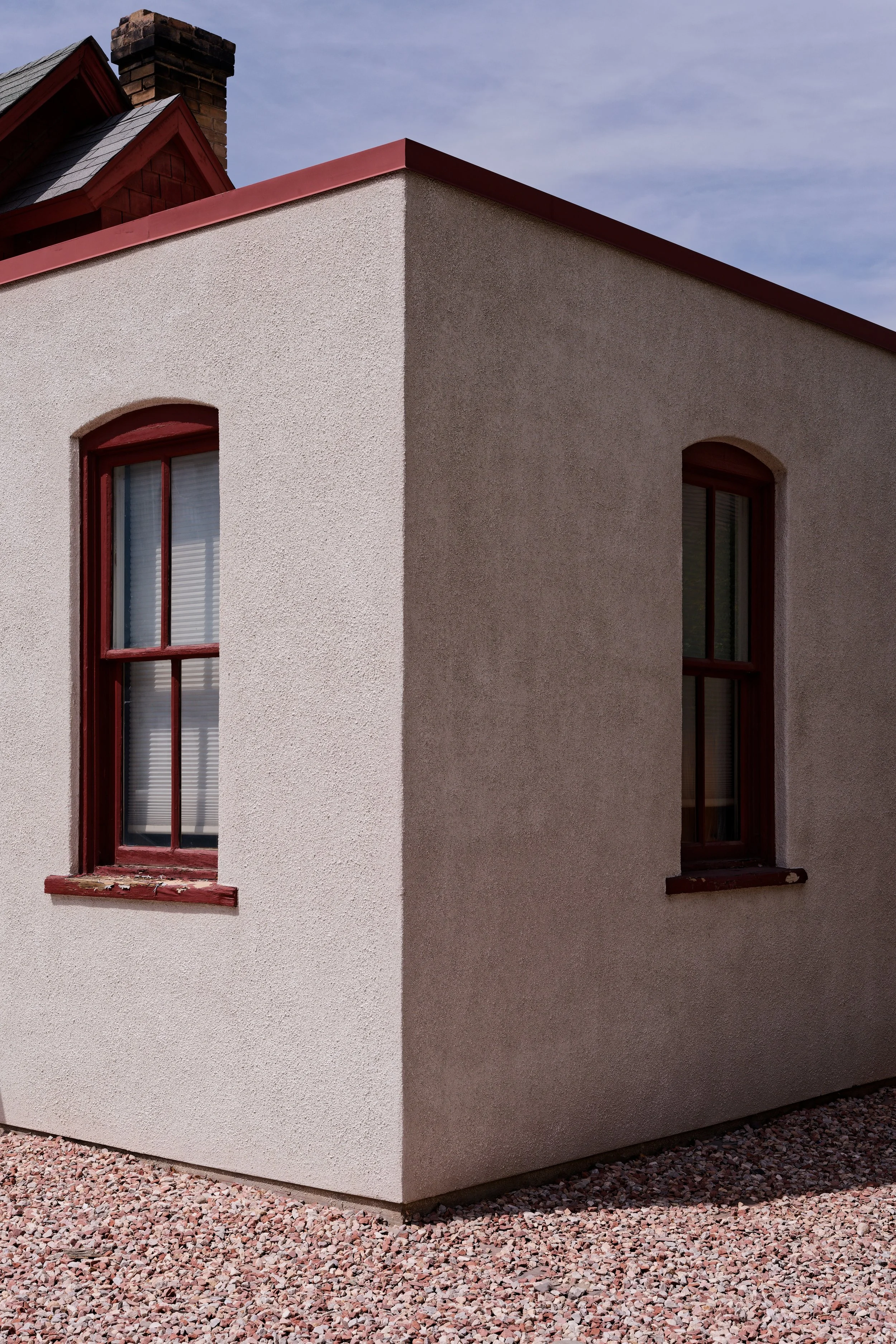 A corner view of a house with two windows with red frames and window sills. The house has a textured white wall on one side and a smooth tan wall on the adjacent side. There is a gravel ground and a chimney visible in the background under a blue sky 